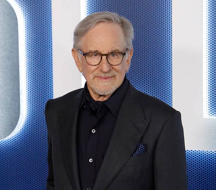 A man in a dark suit and glasses poses confidently at an event, standing in front of a backdrop with large lettering