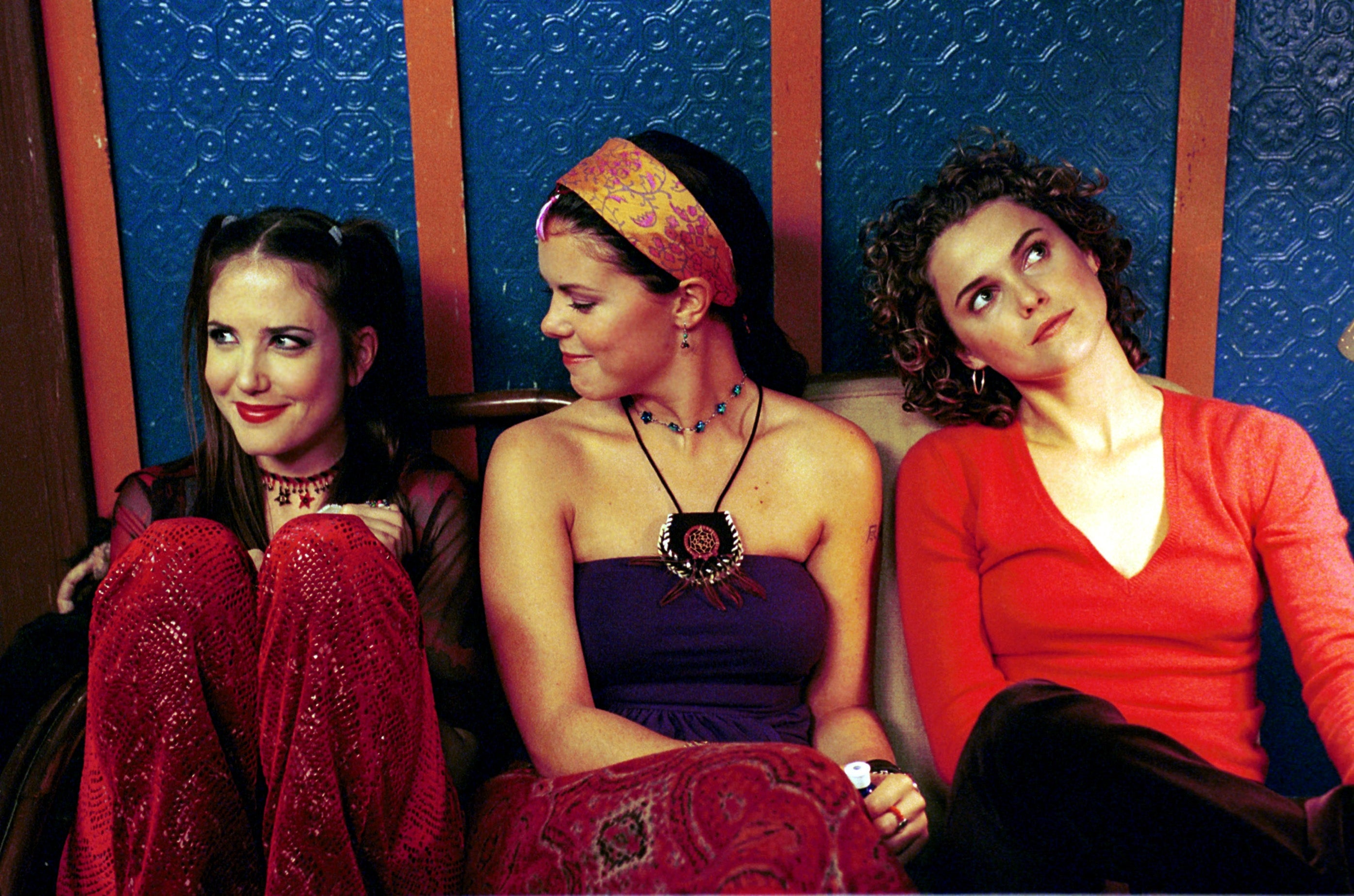 Three women sit together, styled in '90s fashion. One wears a mesh top and patterned pants, another a headband and strapless dress, and the third a V-neck top