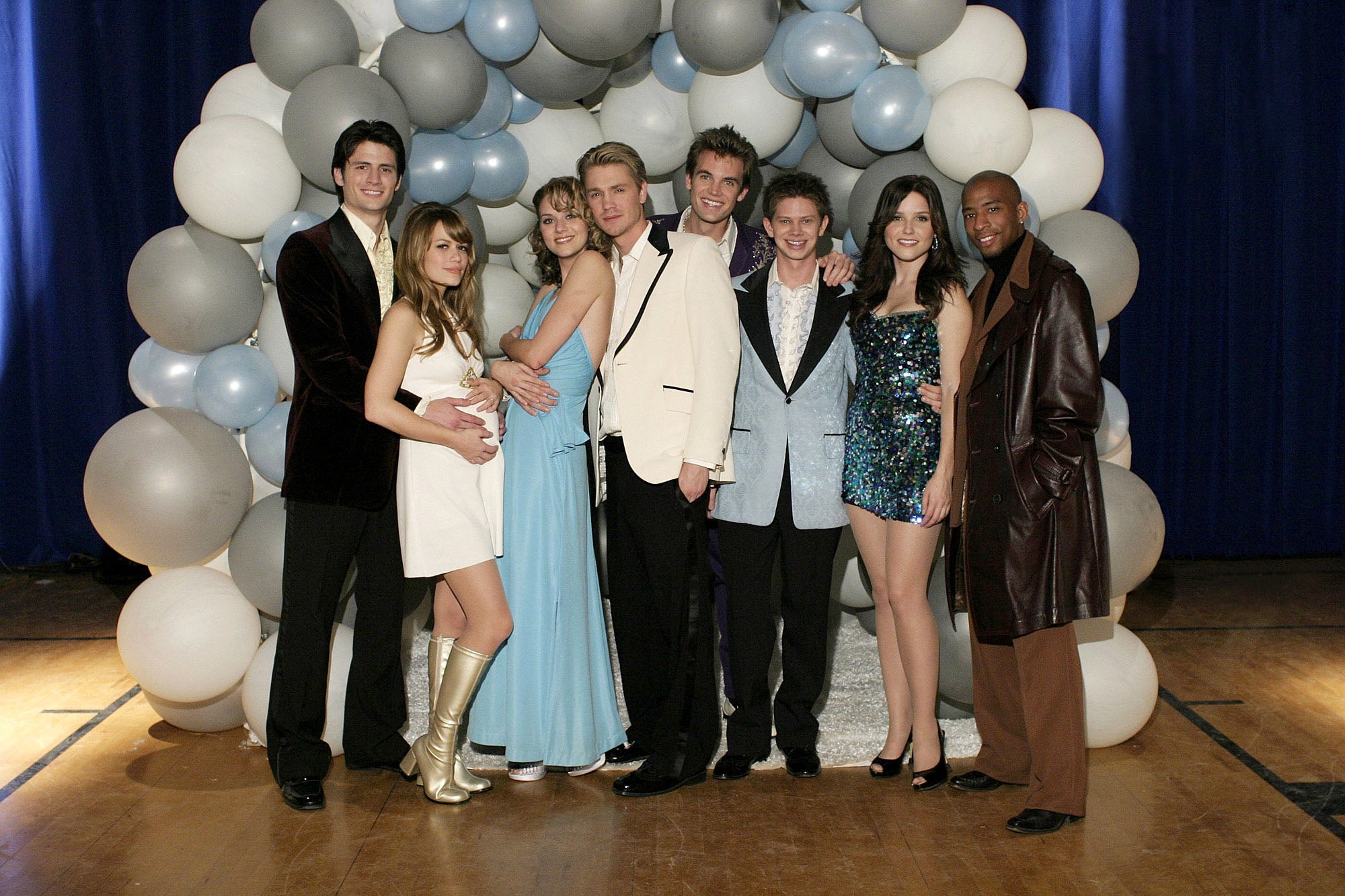 A group of well-dressed people at an event stand in front of a balloon arch. Some wear formal suits; others wear stylish dresses, boots, and a coat