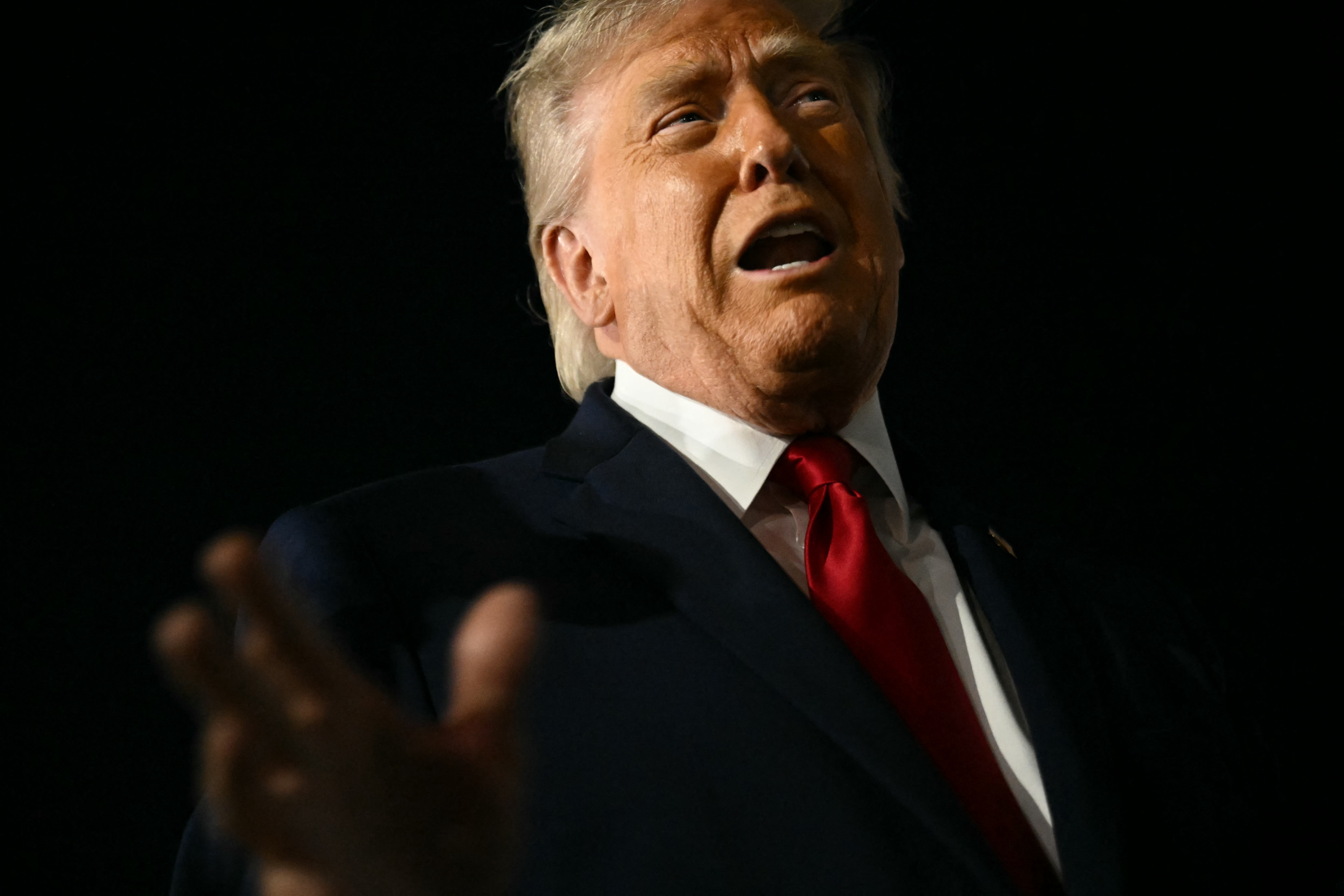 A idiosyncratic successful a suit gestures with 1 hand, appearing to speak, against a acheronian backdrop