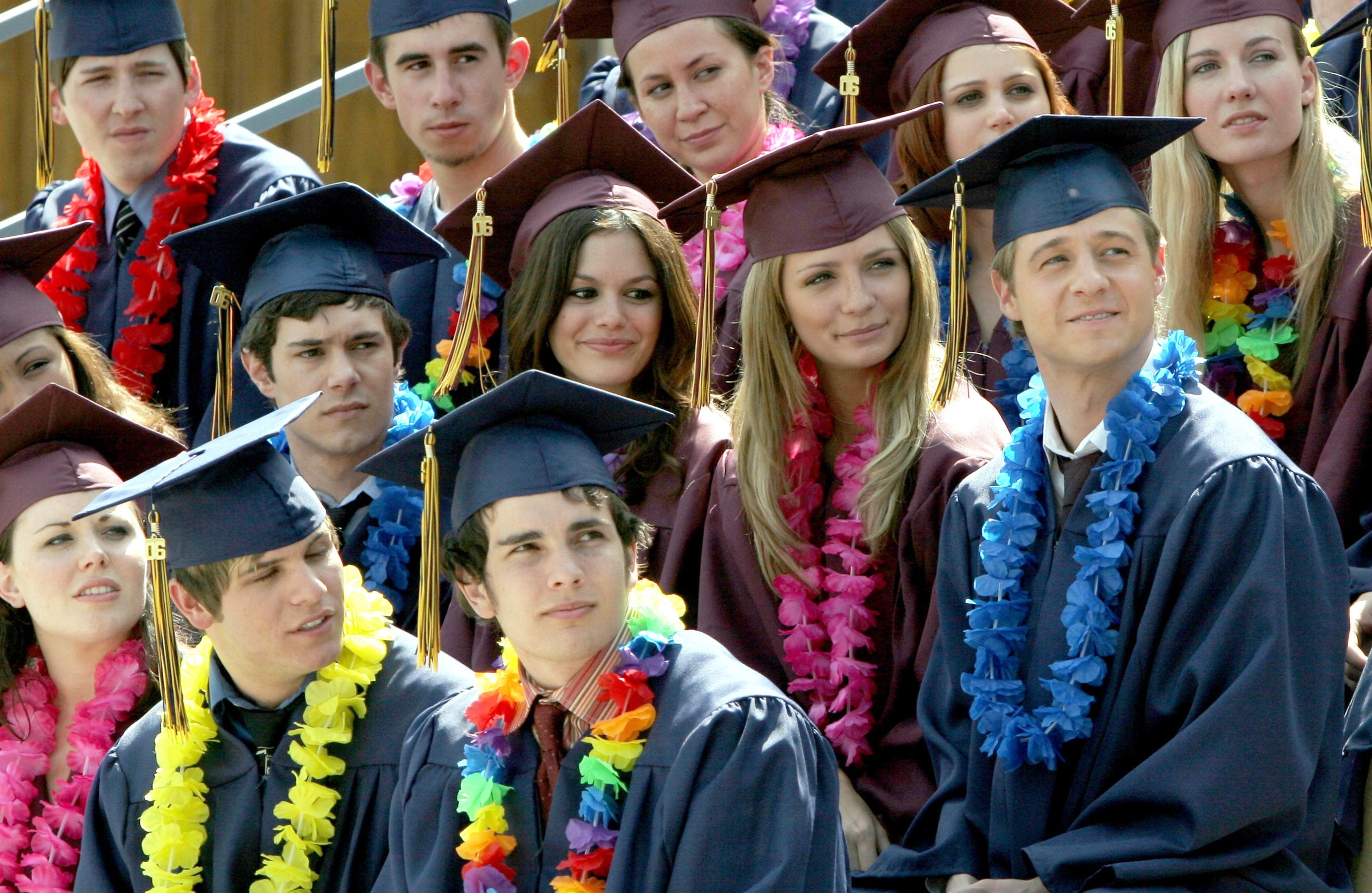 A group of young adults in graduation caps and gowns, adorned with colorful leis, sit together, smiling