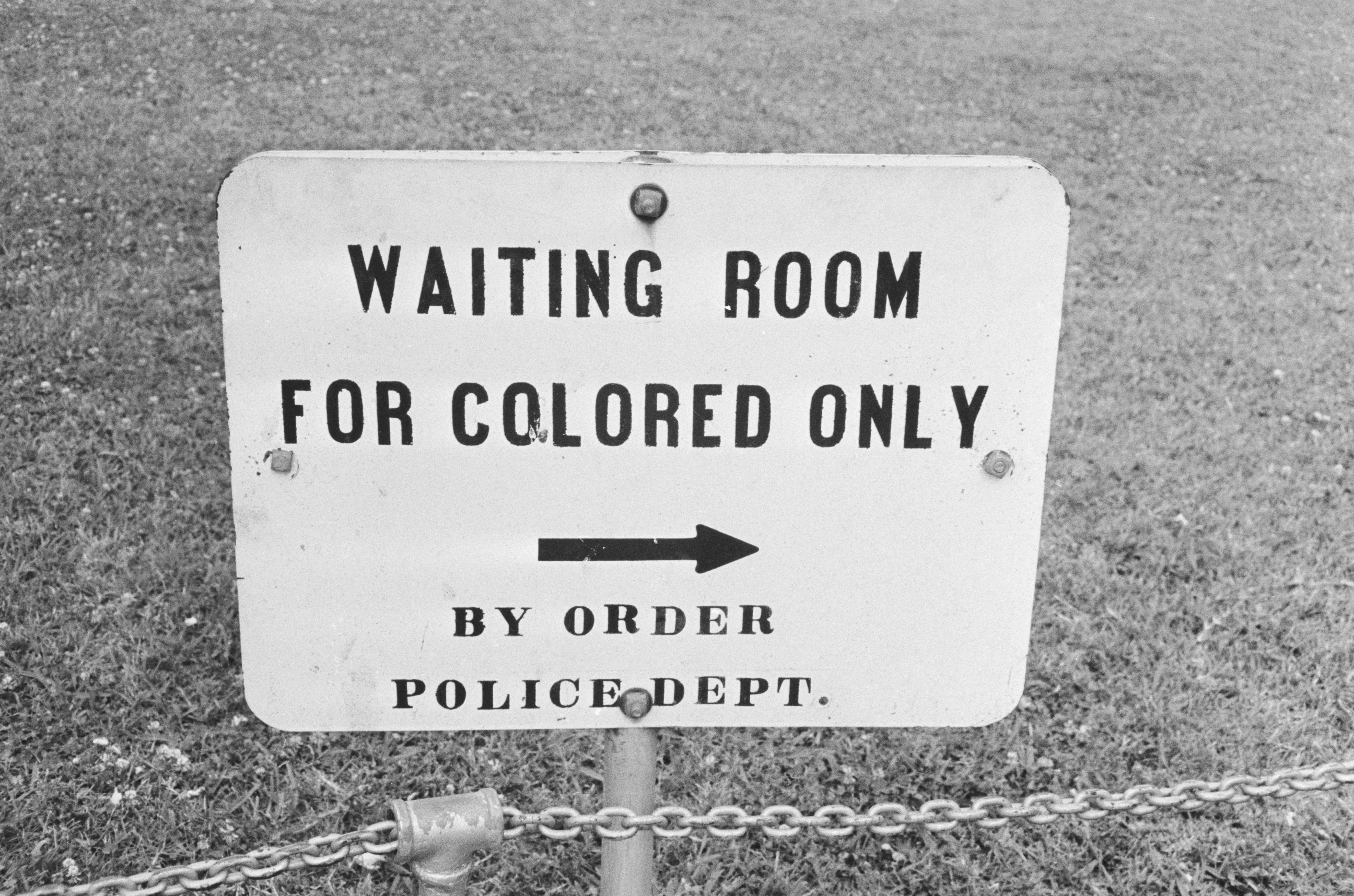 Sign speechmaking "Waiting Room for Colored Only" with an arrow pointing right, followed by "By Order Police Dept." connected a grassy background