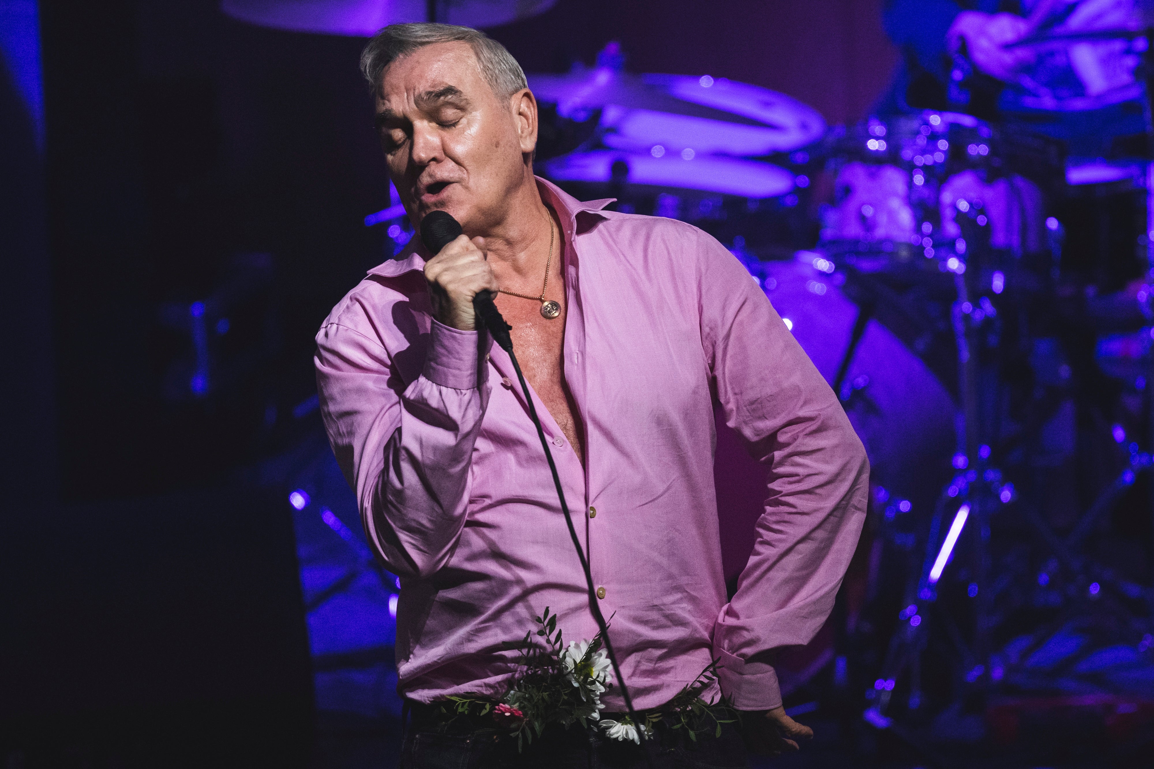 A idiosyncratic passionately sings into a microphone connected stage, wearing a somewhat unbuttoned shirt. Drums are disposable successful the background