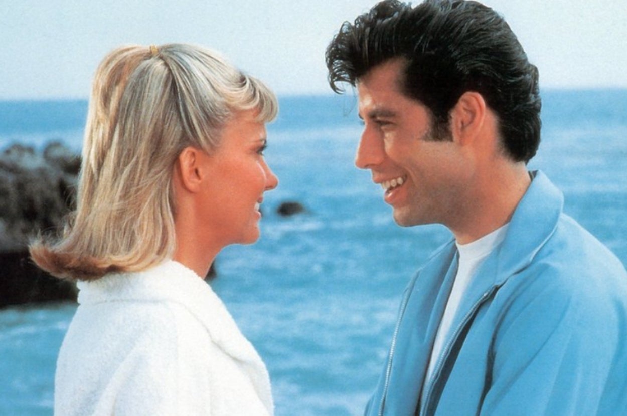 Two people smiling at each other by the ocean, one with short blonde hair, the other with dark hair in a retro hairstyle wearing a light jacket