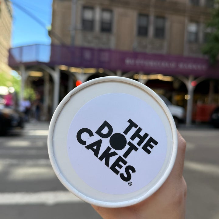 A hand holding a cup labeled "The Dot Cakes" in front of a blurred city street background