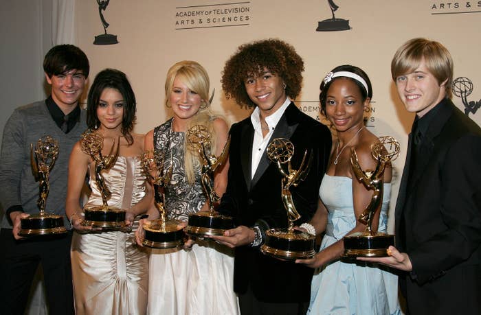The high school musica cast holding emmy awards, dressed in polished formal attire, smiling at an event backdrop