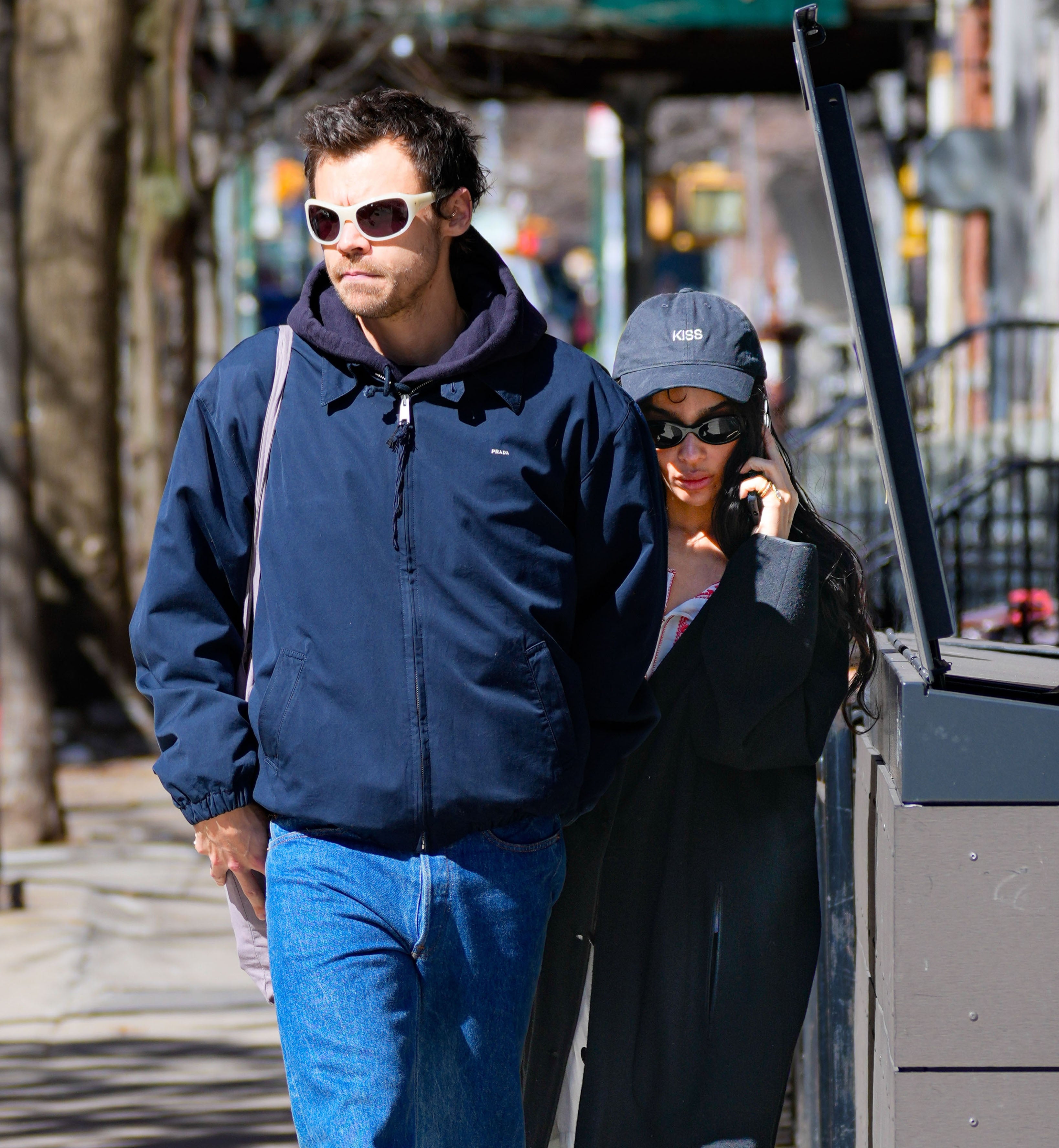 Two radical walking connected a street; 1 wears a overgarment and sunglasses, the different wears a headdress and is talking connected the phone