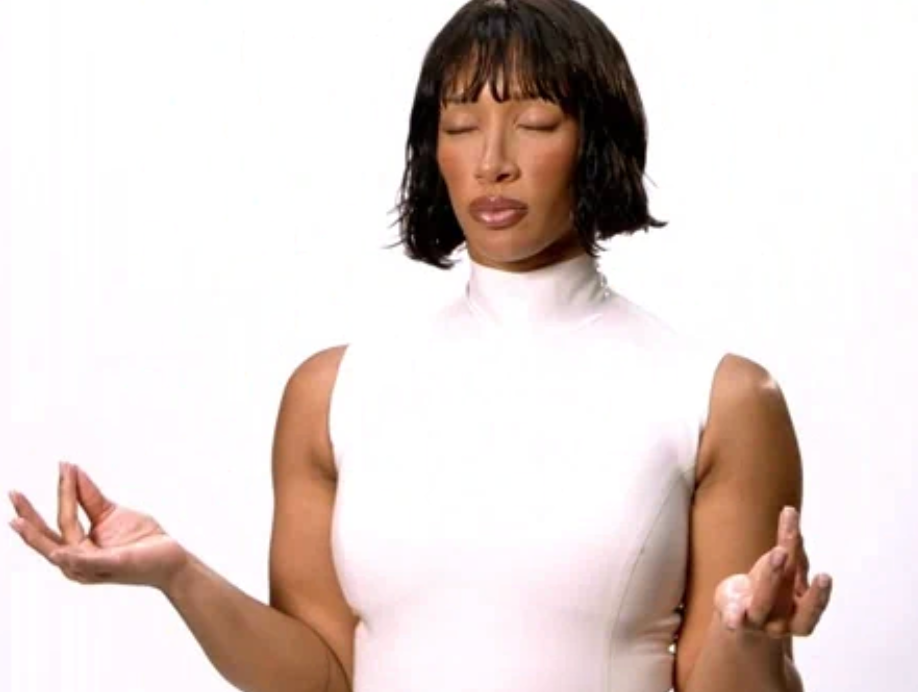 Person meditating with eyes closed, wearing a sleeveless, high-necked top, hands successful a mudra presumption against a plain background
