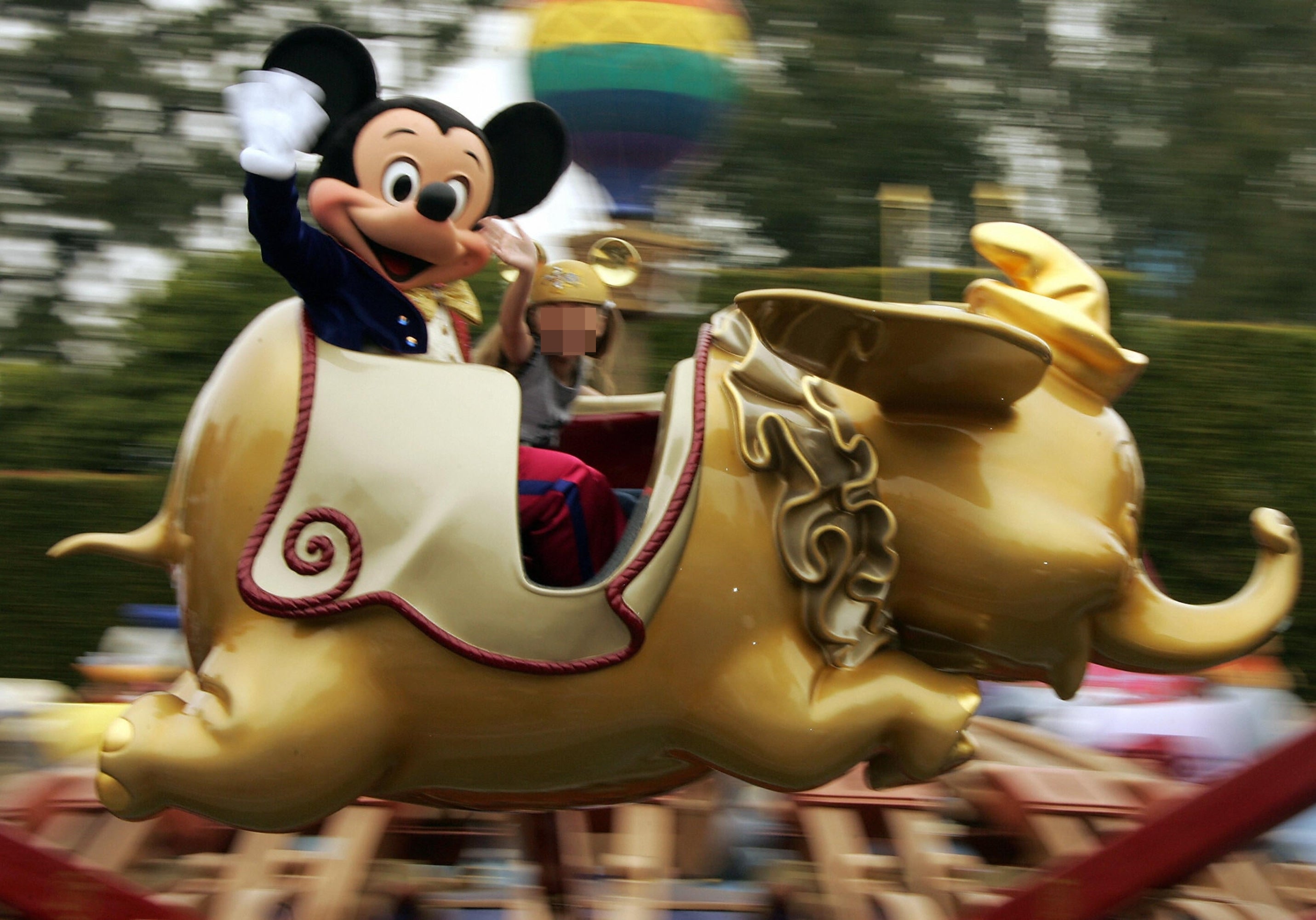 Mickey Mouse waves portion riding a Dumbo elephant thrust with a smiling kid sitting beside him