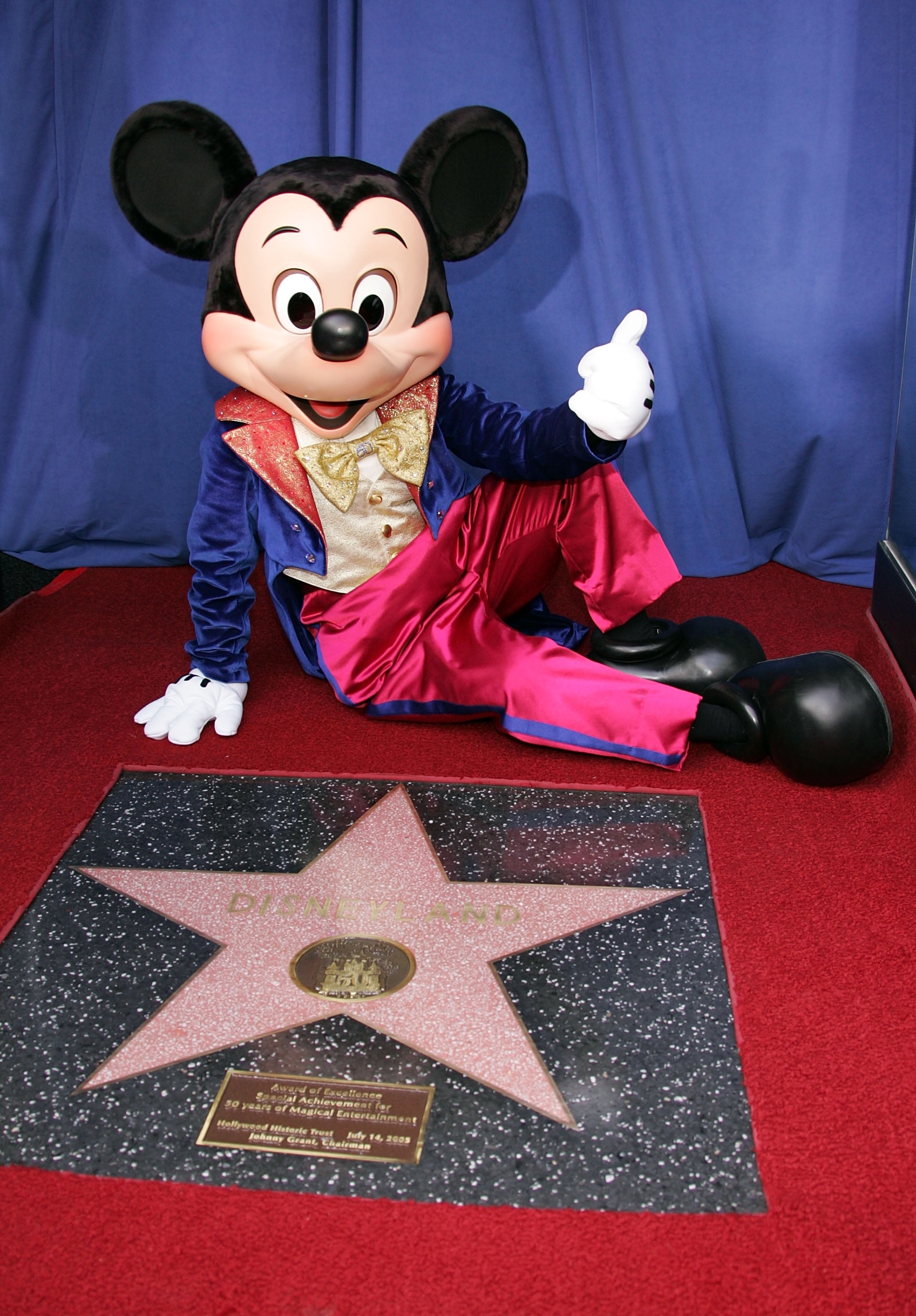 Mickey Mouse successful a colorful outfit sits connected a reddish carpet adjacent to a Hollywood Walk of Fame star, giving a thumbs-up