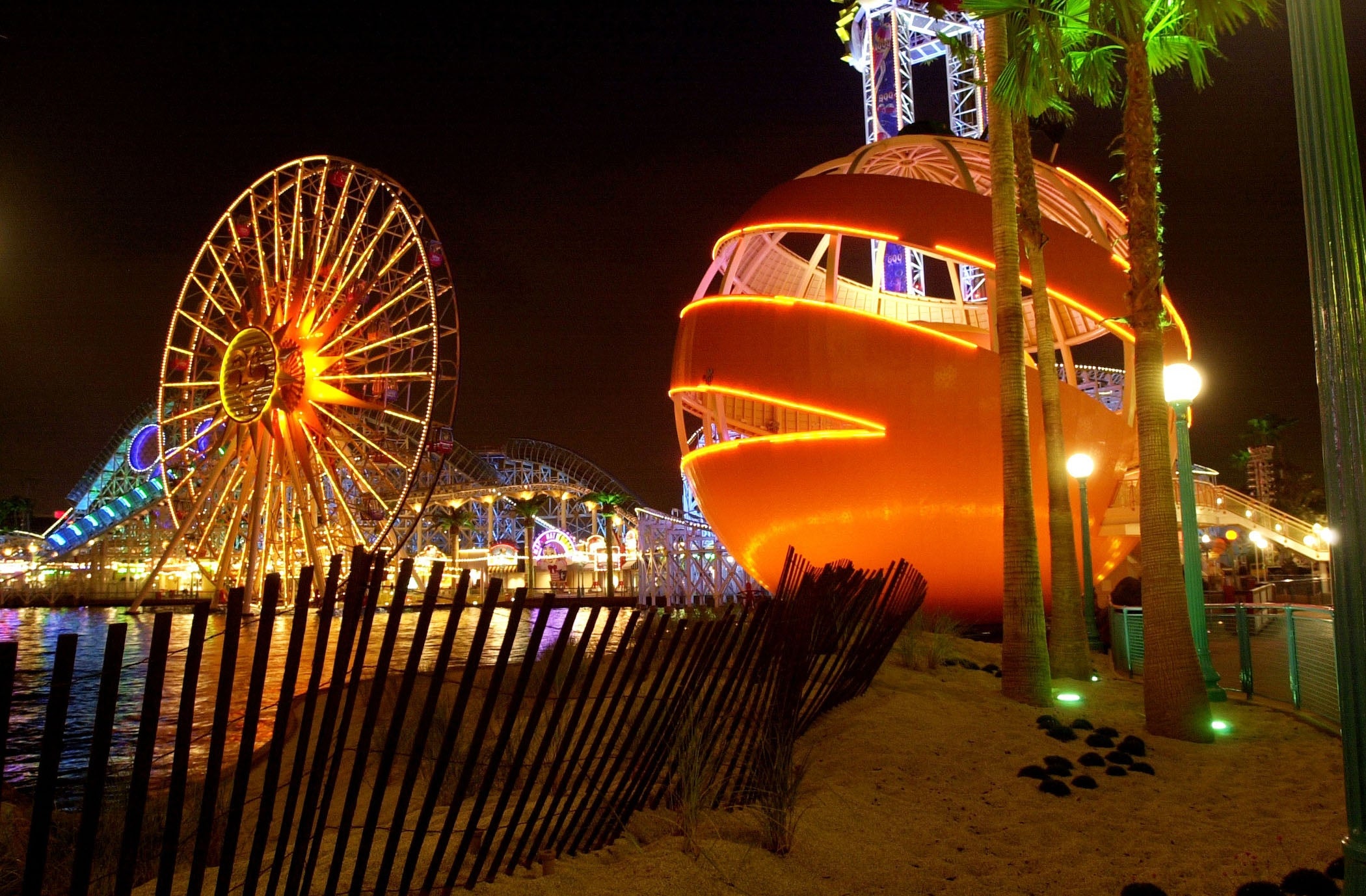 Amusement parkland astatine nighttime with a ample Ferris instrumentality and an orange, spherical structure. Palm trees and soil heighten the lively atmosphere