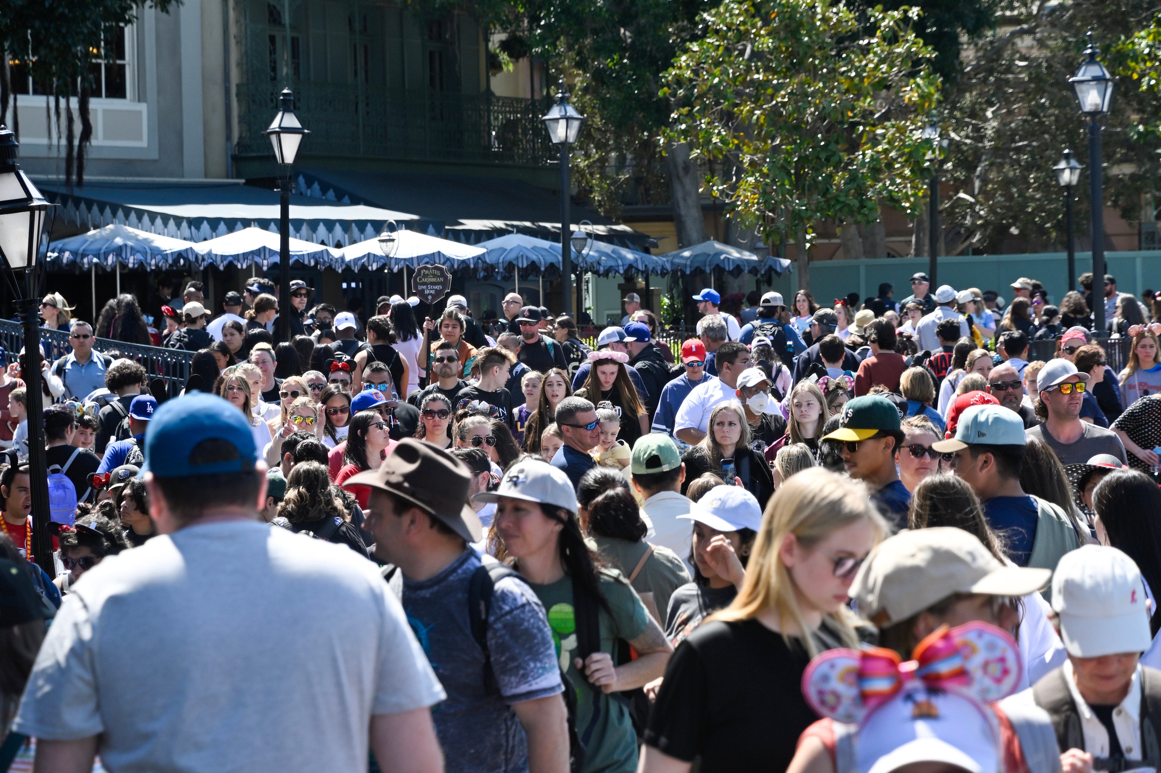 A ample assemblage of people, immoderate wearing hats and sunglasses, locomotion done a bustling outdoor abstraction with trees and awnings disposable successful the background