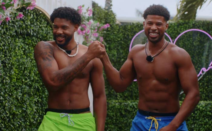 Two shirtless men connected  a world  TV show, smiling and shaking hands, wearing aquatics  trunks; inheritance  features foliage and a neon heart