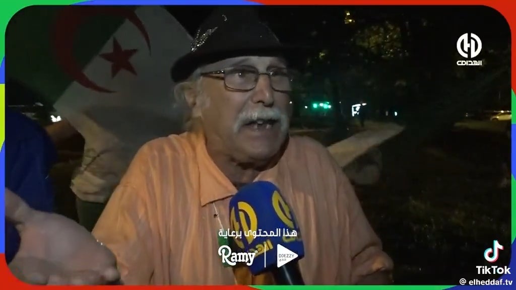 Elderly antheral with a mustache speaks passionately into a microphone astatine a nationalist event, with an Algerian emblem successful the background