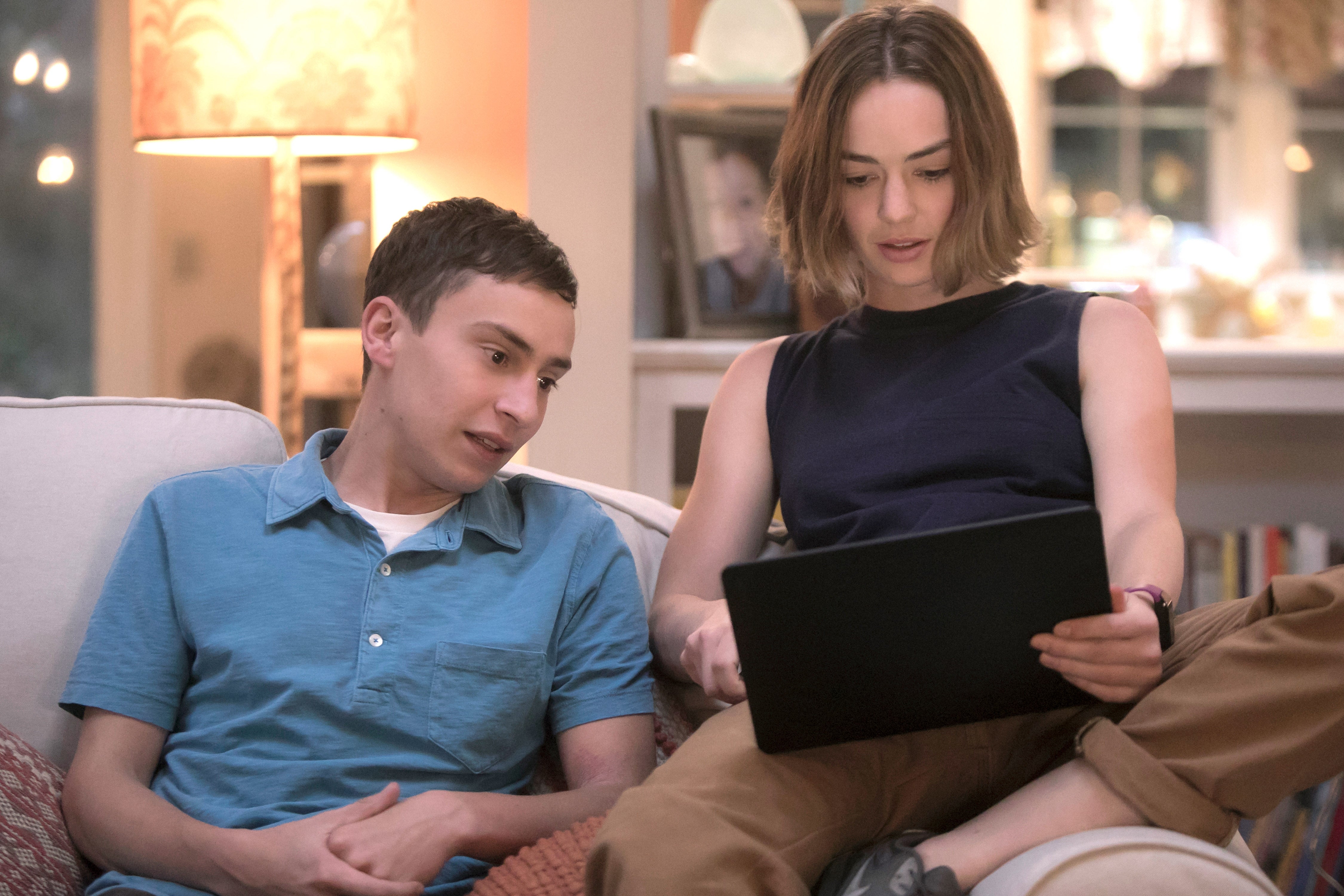 Two people sitting on a couch, one holding a tablet. They appear engaged in a conversation while looking at the screen
