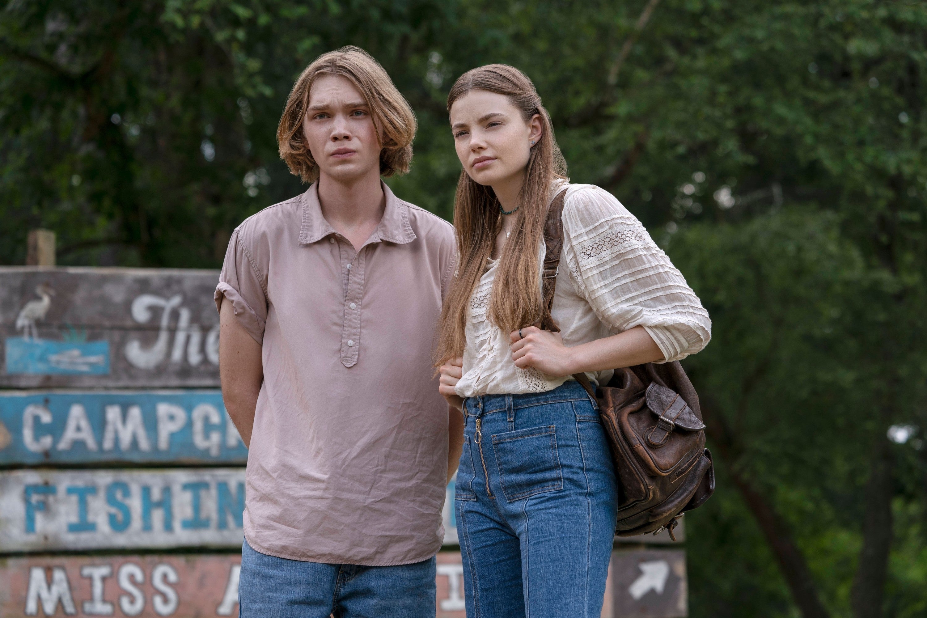 Two people stand outdoors near a rustic sign. One wears a short-sleeve shirt; the other, a long-sleeve top and jeans, holding a backpack