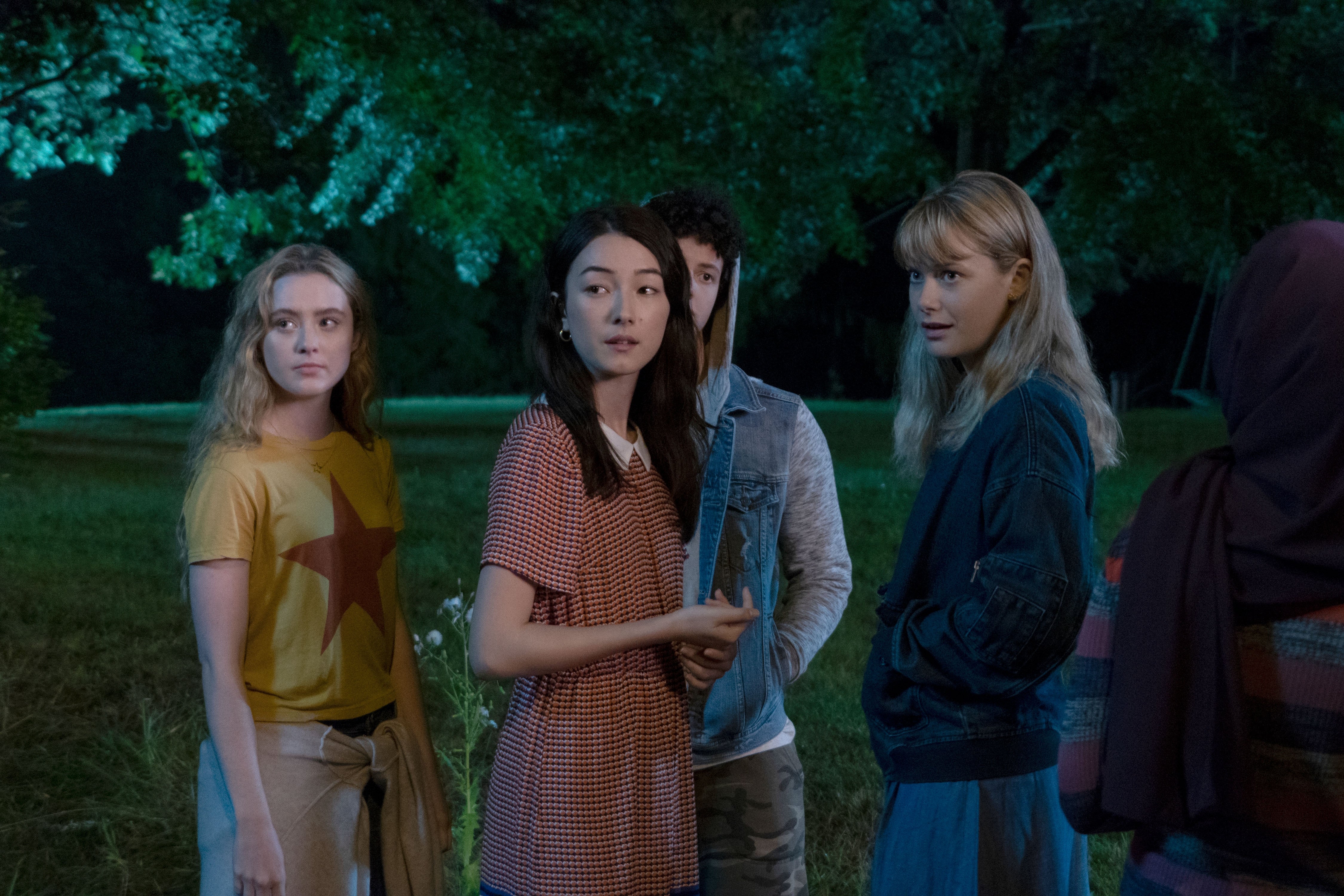 A group of young adults stands outdoors at night, appearing concerned, as they listen intently