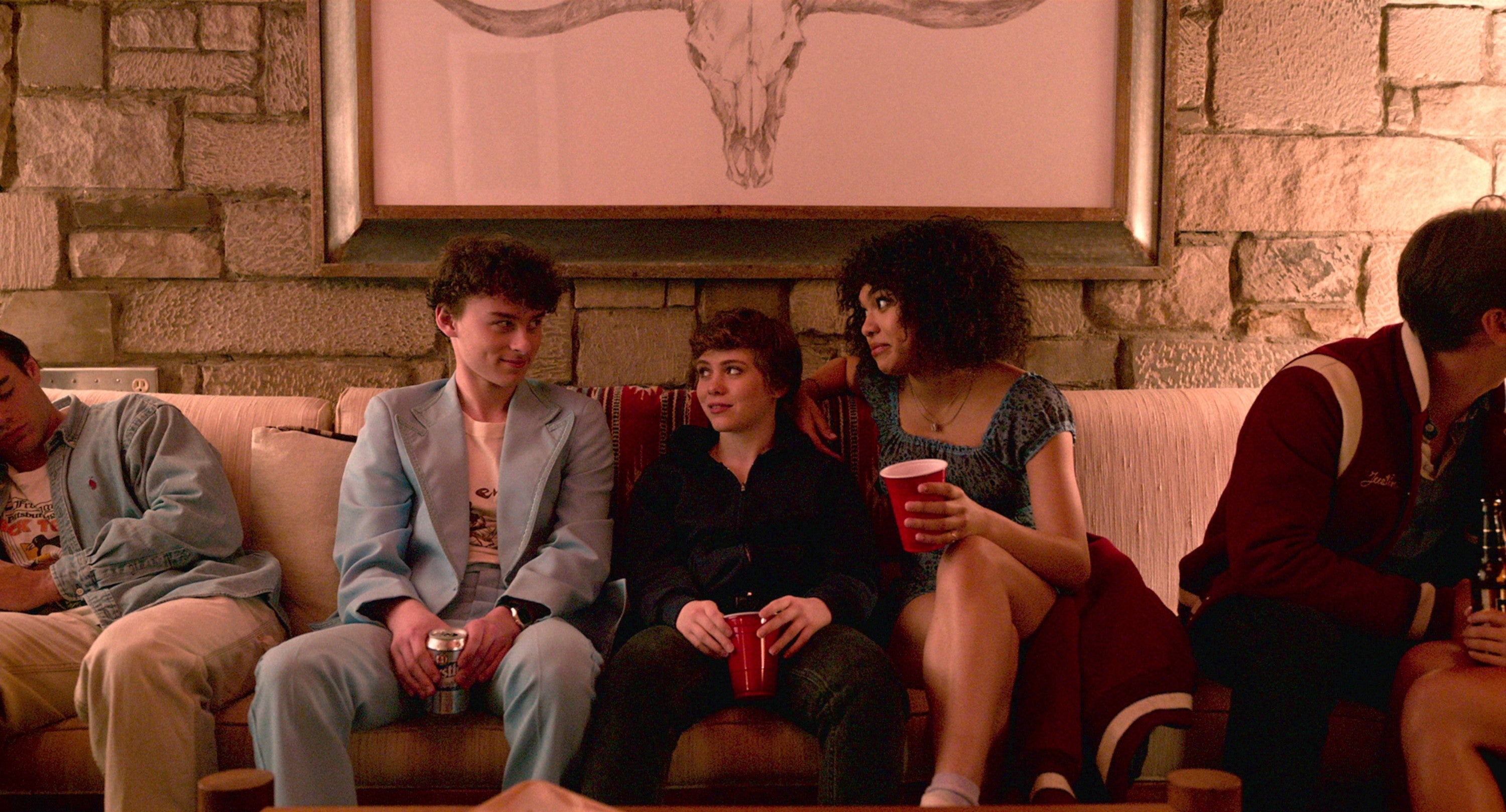Three people are sitting on a couch at a casual party, holding red cups and talking under wall art featuring an animal skull