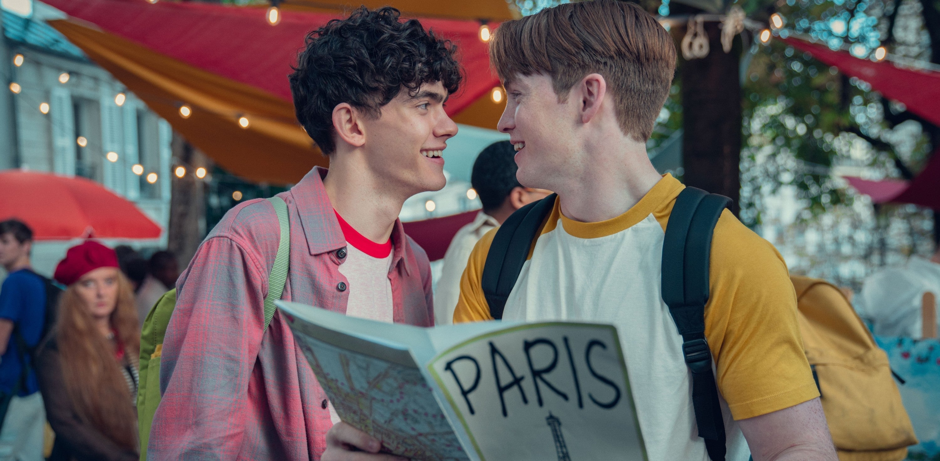 Two smiling young men stand close, one holding a "Paris" map in a lively outdoor setting with string lights