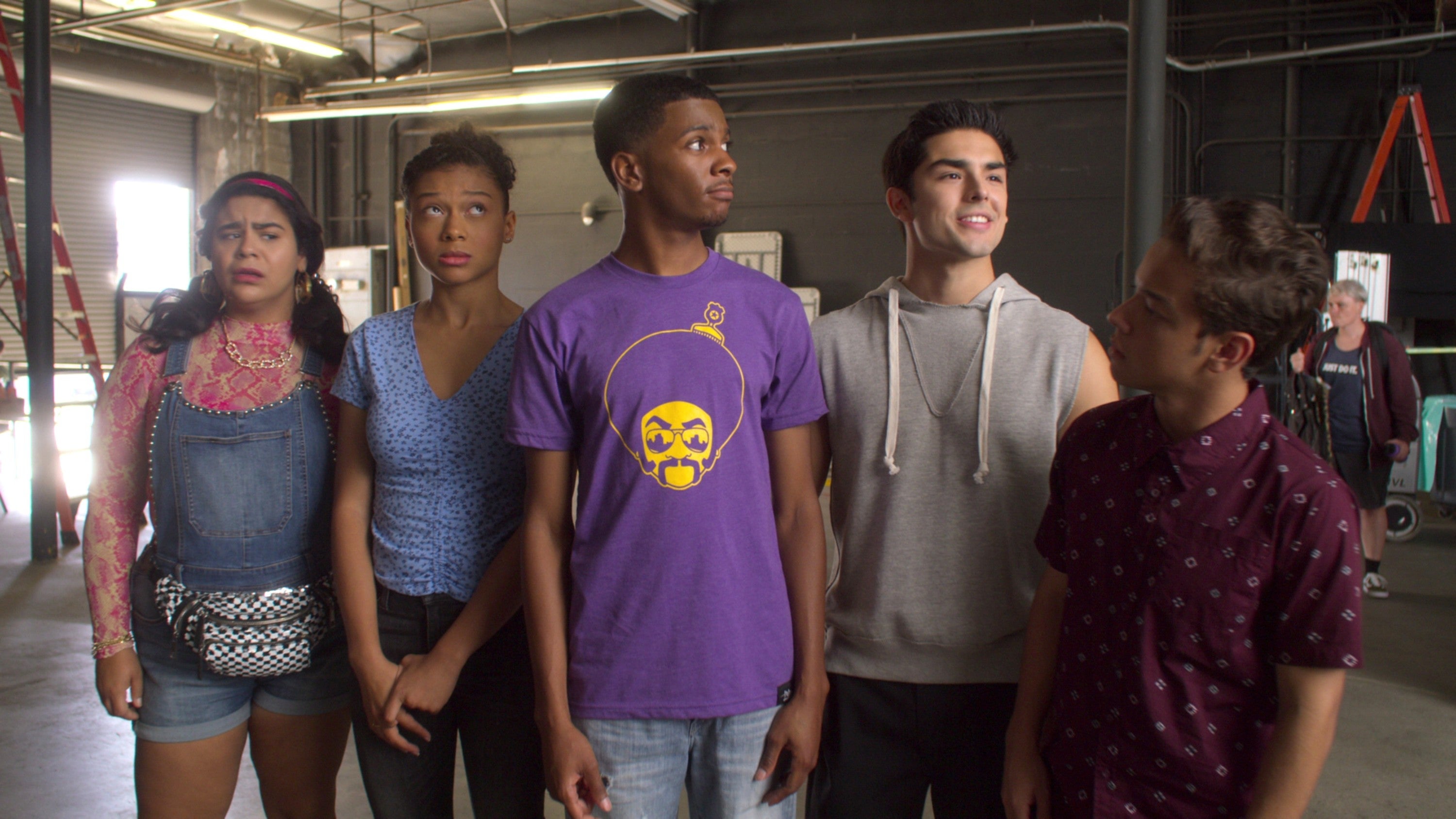 Five people stand in a warehouse, looking curious. One wears a purple graphic tee, another dons denim overalls, while the others wear casual outfits