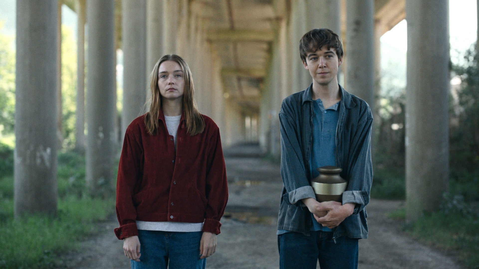 Two young people stand under a bridge. One holds an urn, and both wear casual attire, looking straight ahead