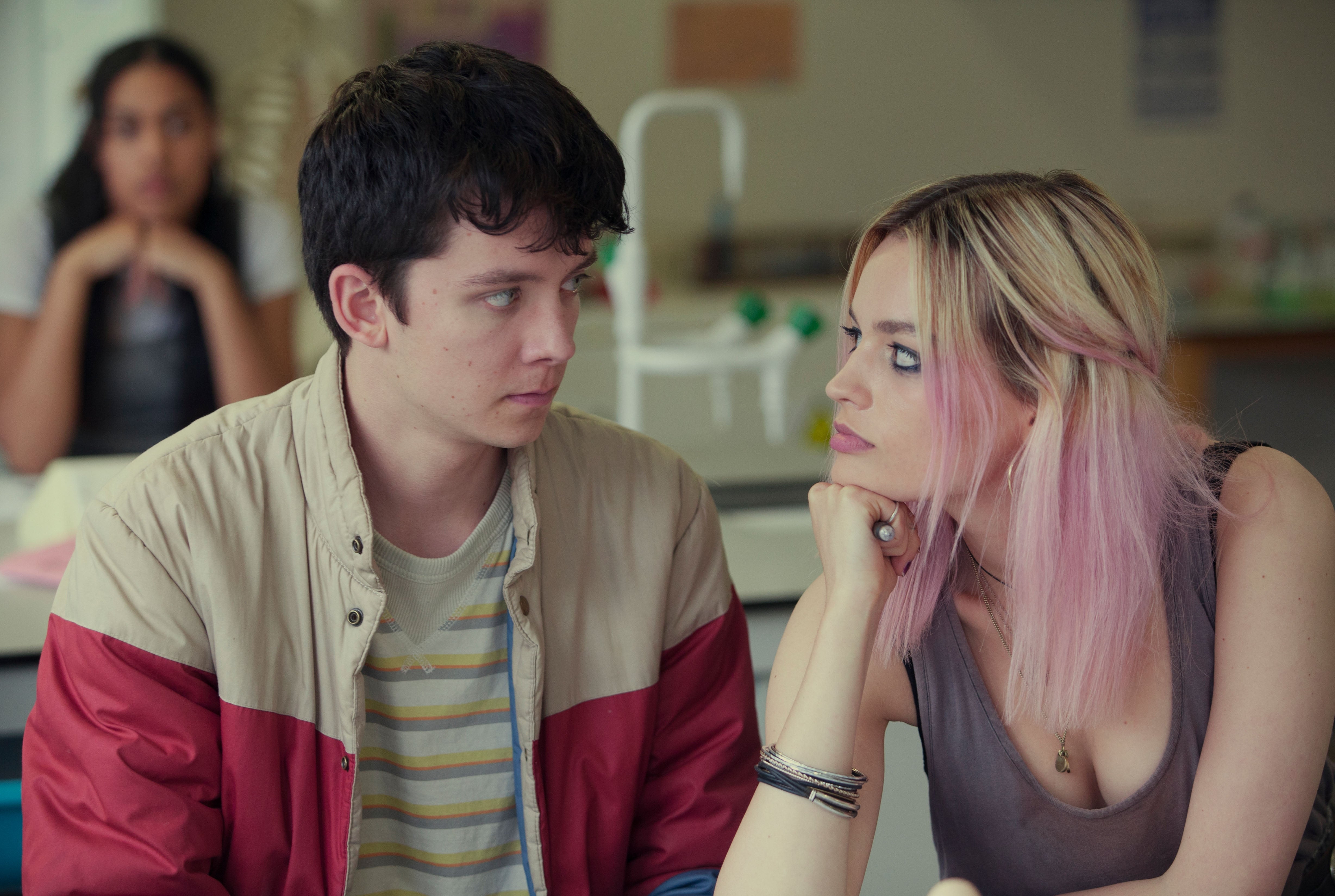 Two people sit at a table, one gazing intently at the other. The setting appears to be casual, possibly a school or similar environment