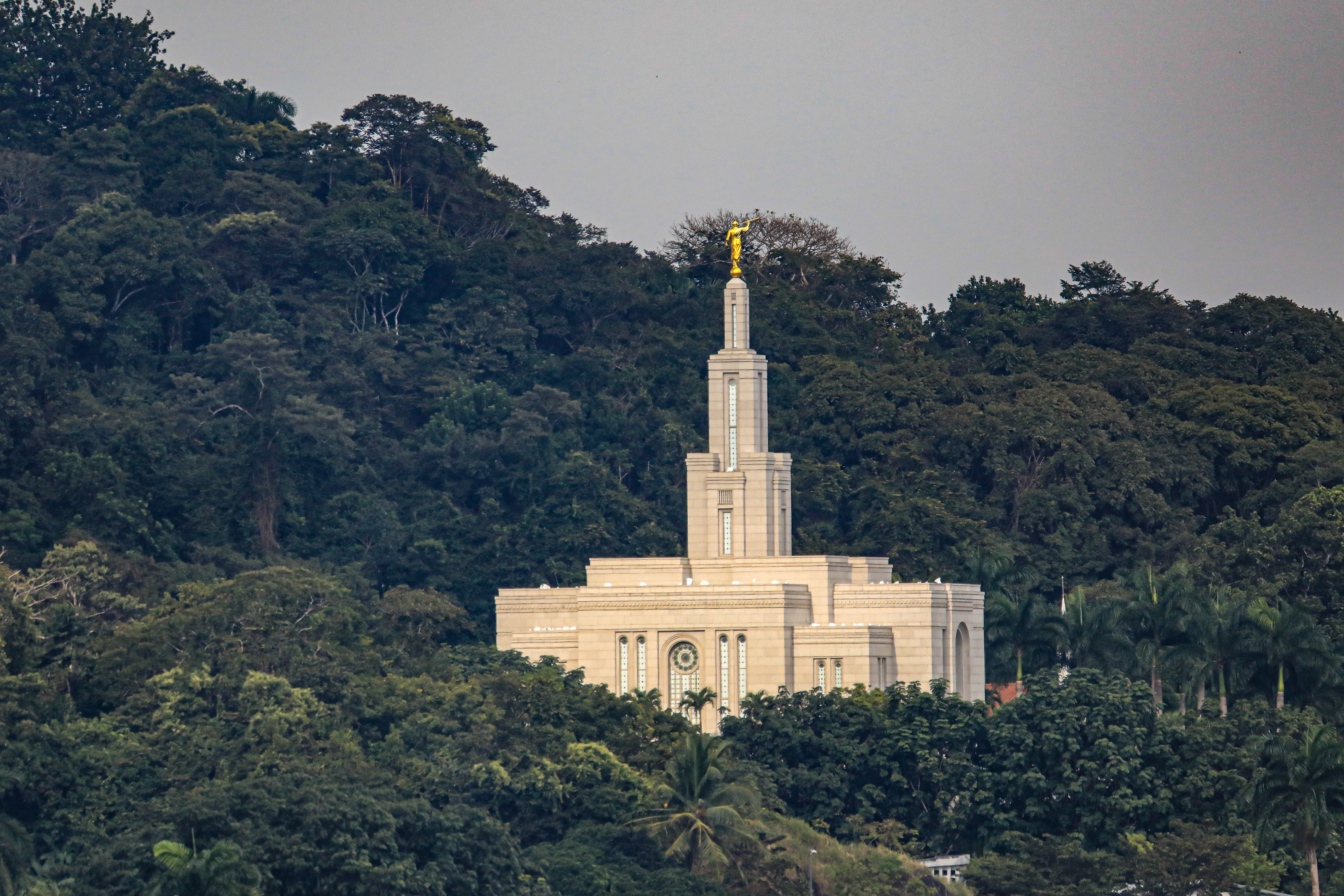 A temple with a gangly spire stands surrounded by lush greenish trees connected a hillside