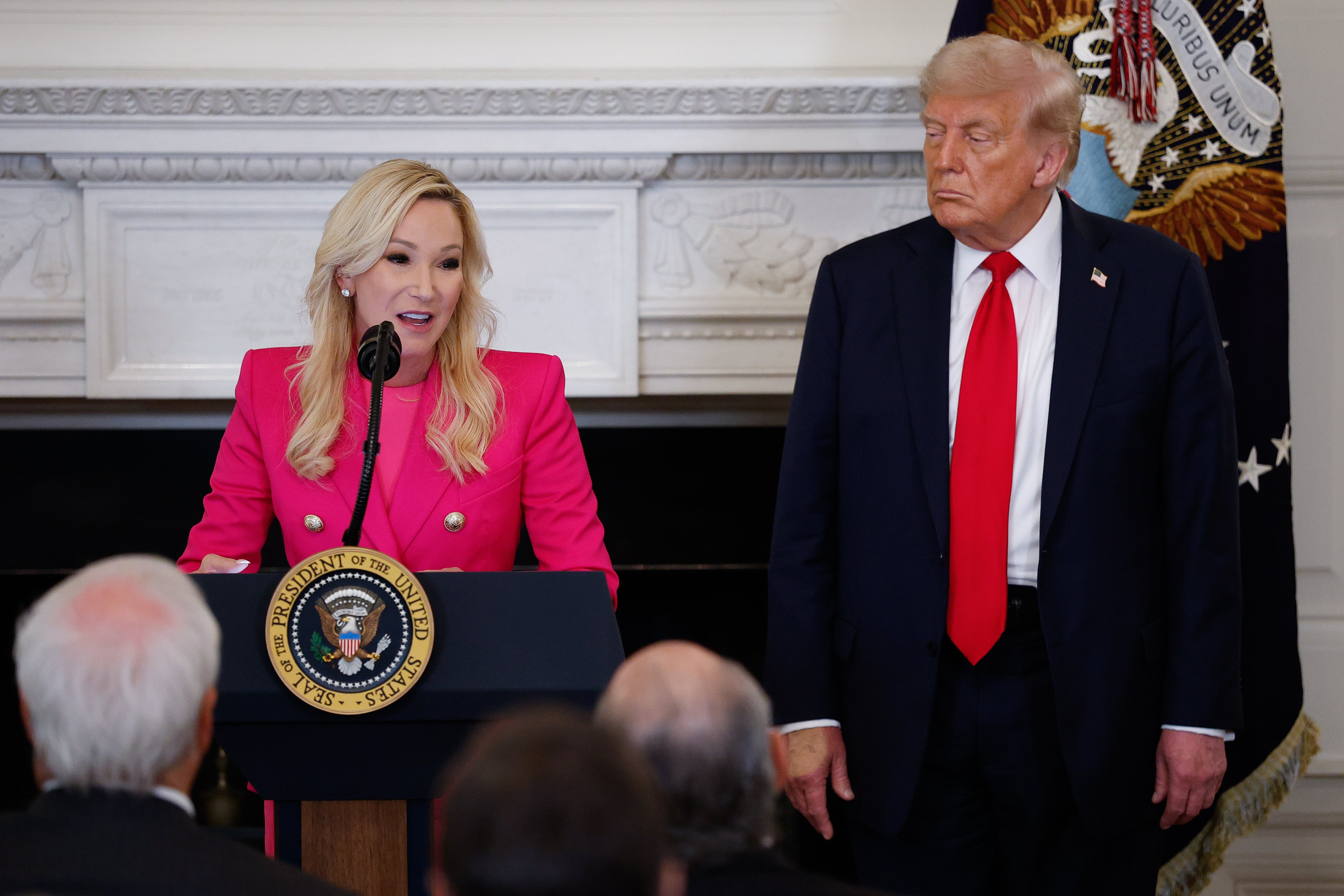 A pistillate successful a nonrecreational suit speaks astatine a podium with a statesmanlike seal, portion a antheral successful a suit and reddish necktie stands beside her successful a ceremonial setting