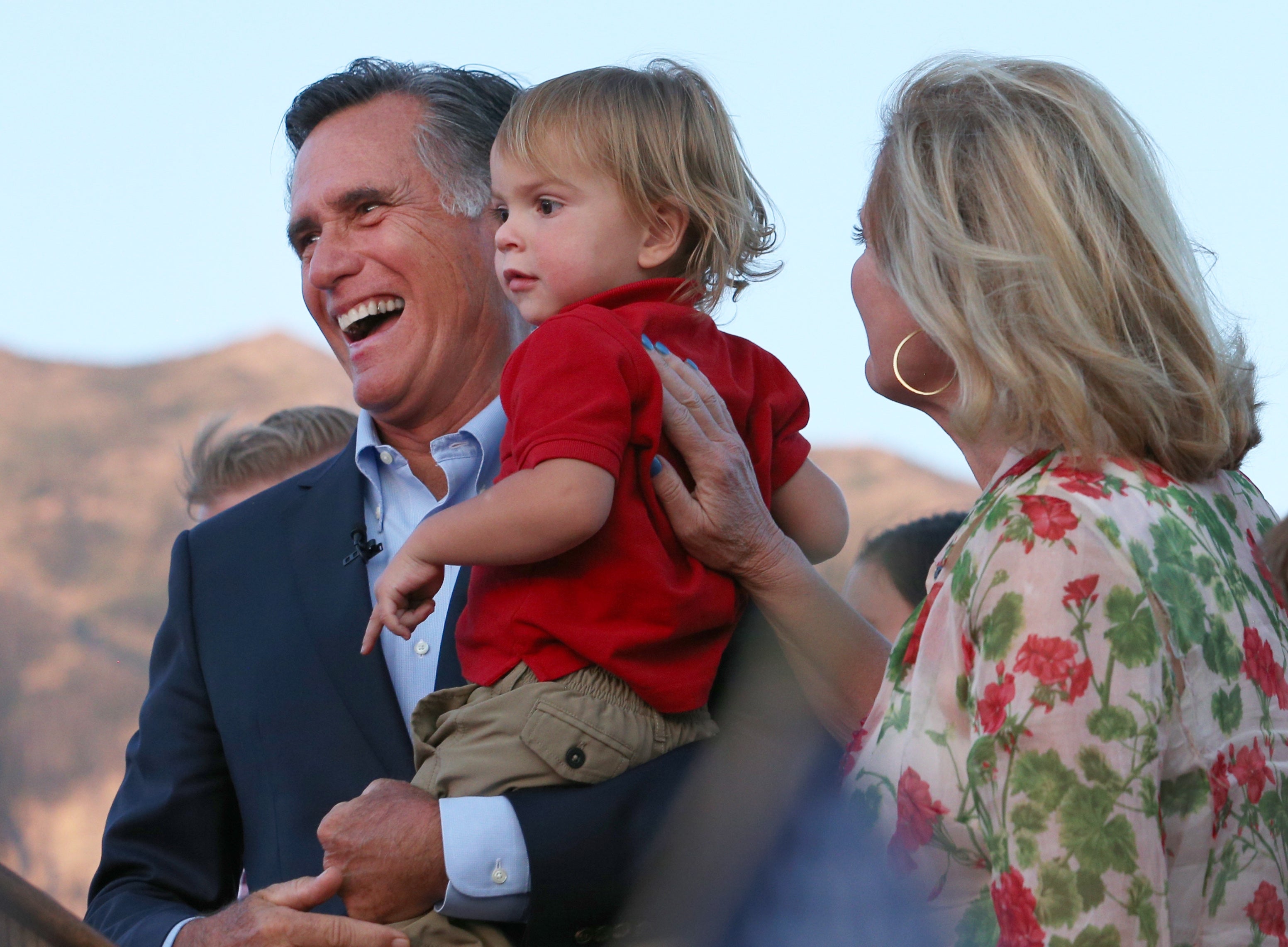 A idiosyncratic successful a suit smiles portion holding a young child. Another idiosyncratic successful floral attire stands beside them, facing a scenic backdrop