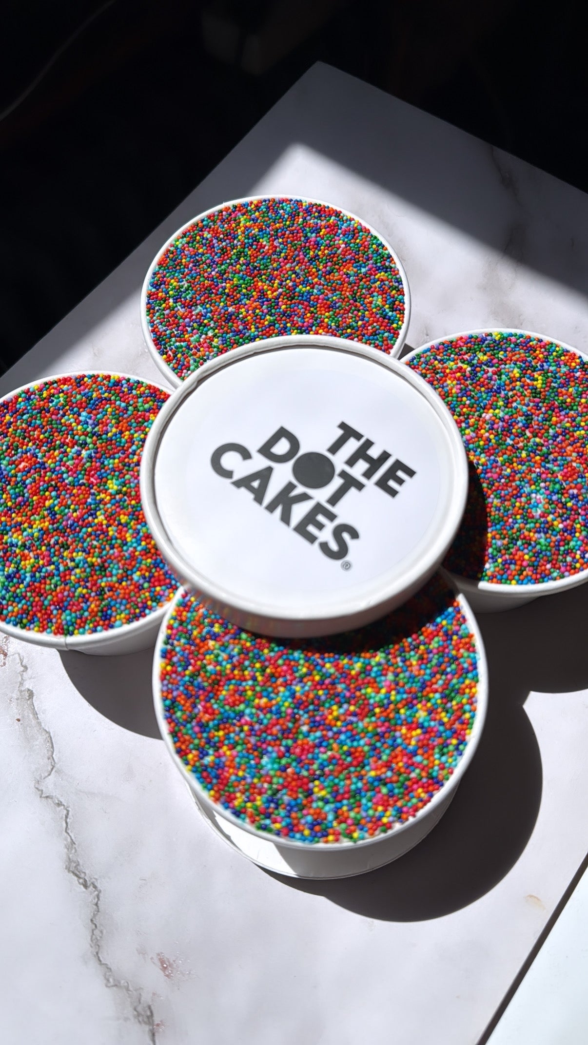 Containers of colorful sprinkles surround a central container labeled "The Dot Cakes" on a marble surface