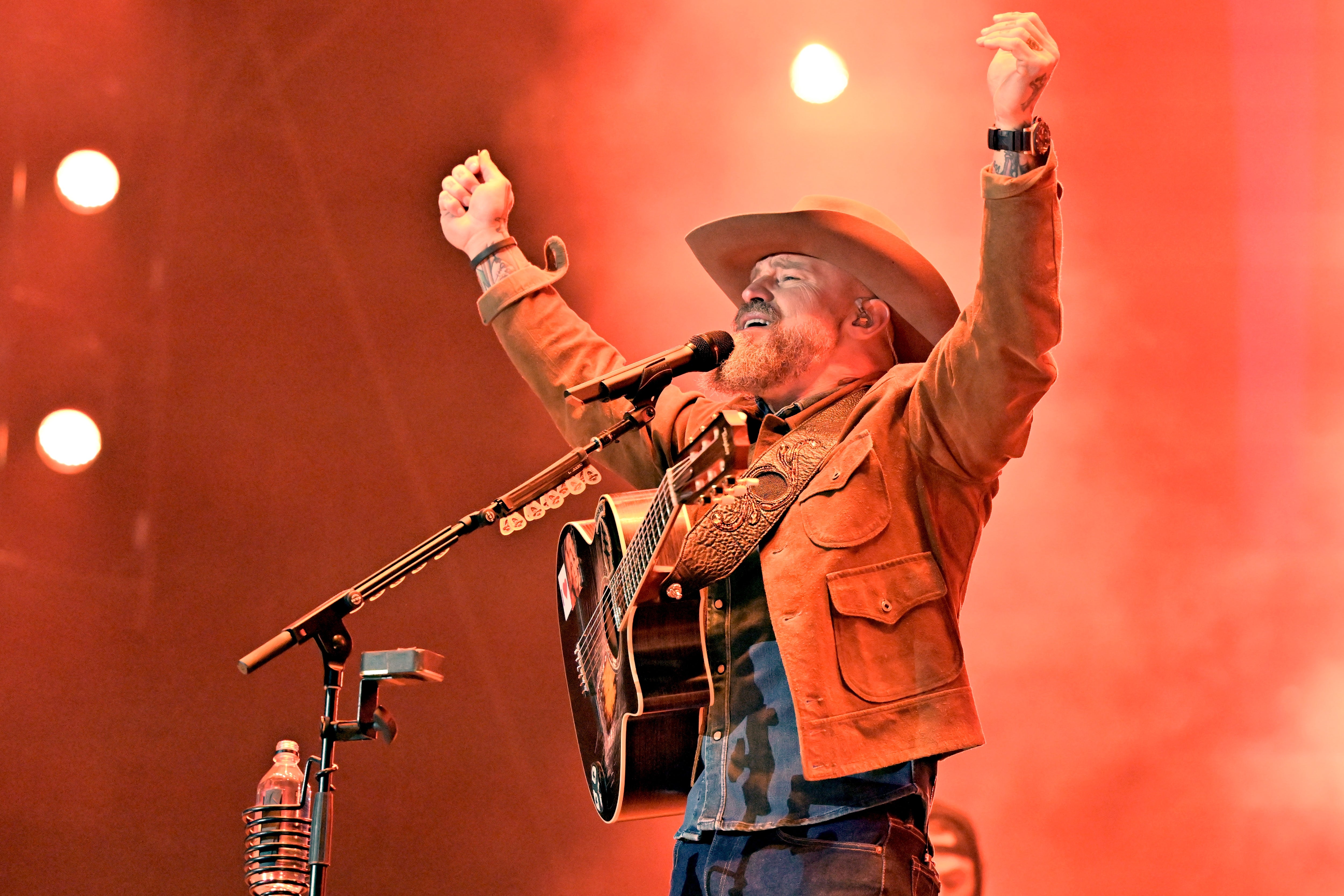 Country instrumentalist performs energetically connected stage, wearing a cowboy chapeau and leather jacket, raising some arms successful excitement