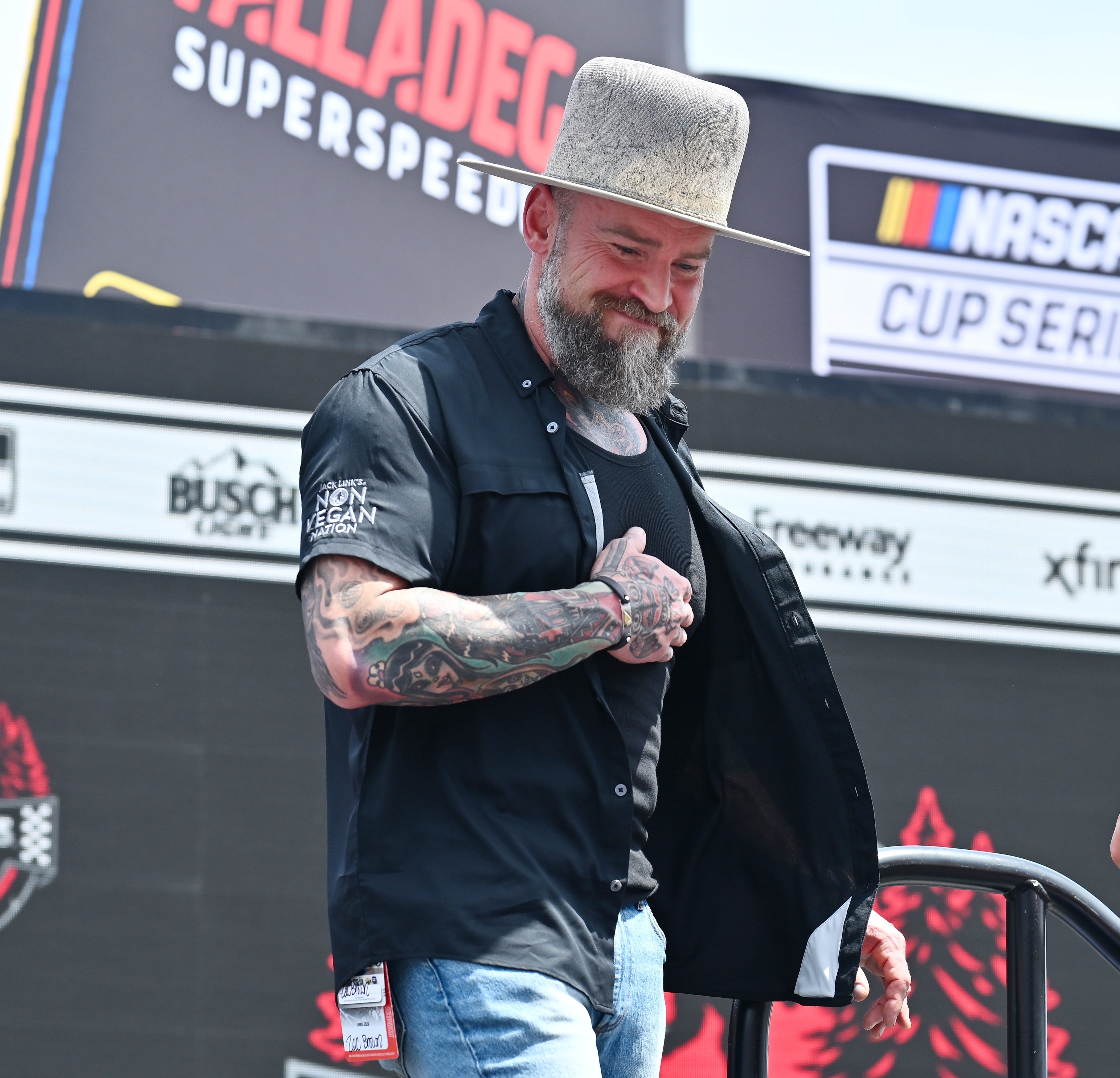 Person with a bushy beard wearing a wide-brimmed hat, achromatic short-sleeved shirt, and jeans, connected signifier astatine NASCAR event