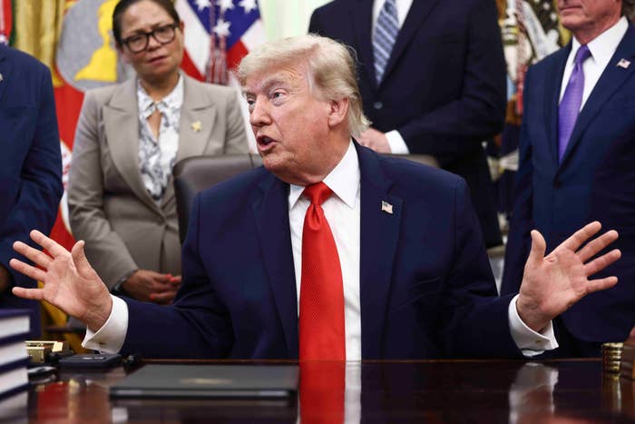 A antheral   successful  a suit   with a reddish  necktie  is gesturing with his hands portion    sitting astatine  a desk, surrounded by radical   successful  a ceremonial  setting