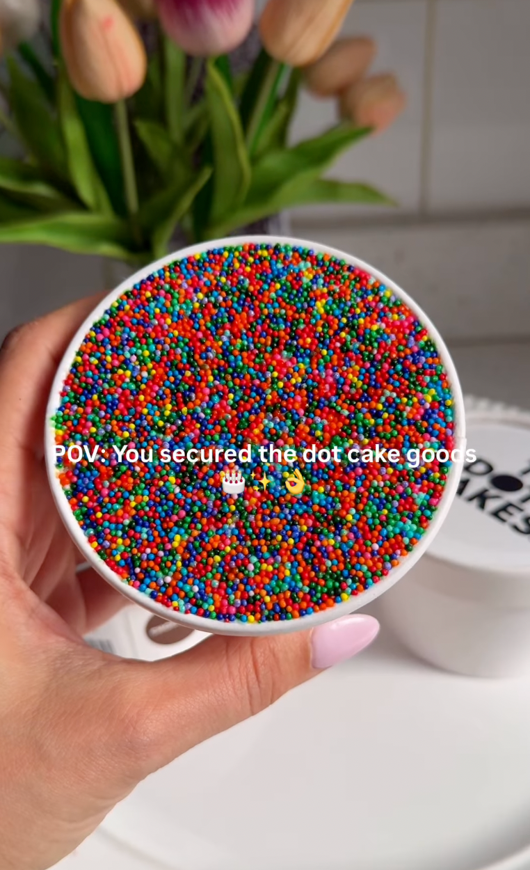 A hand holds a bowl filled with multicolored sprinkles. Text overlay: "POV: you secured the dot cake goods."