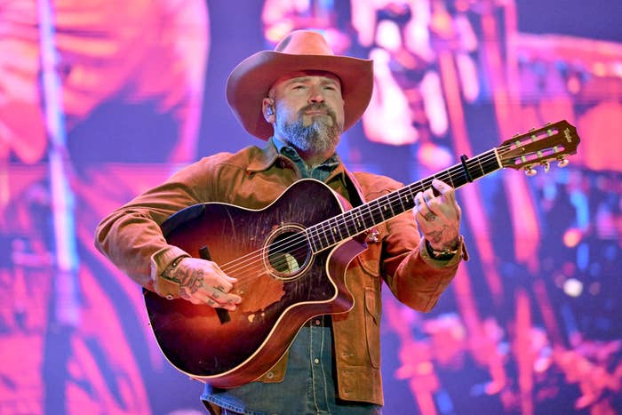 A instrumentalist  performs connected  signifier    wearing a cowboy hat, a jacket, and holding an acoustic guitar
