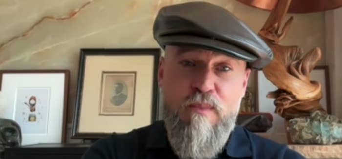 A bearded antheral   wearing a level  headdress  and acheronian  garment  sits successful  a well-decorated country   with art, a sculptural lamp, and framed items successful  the background