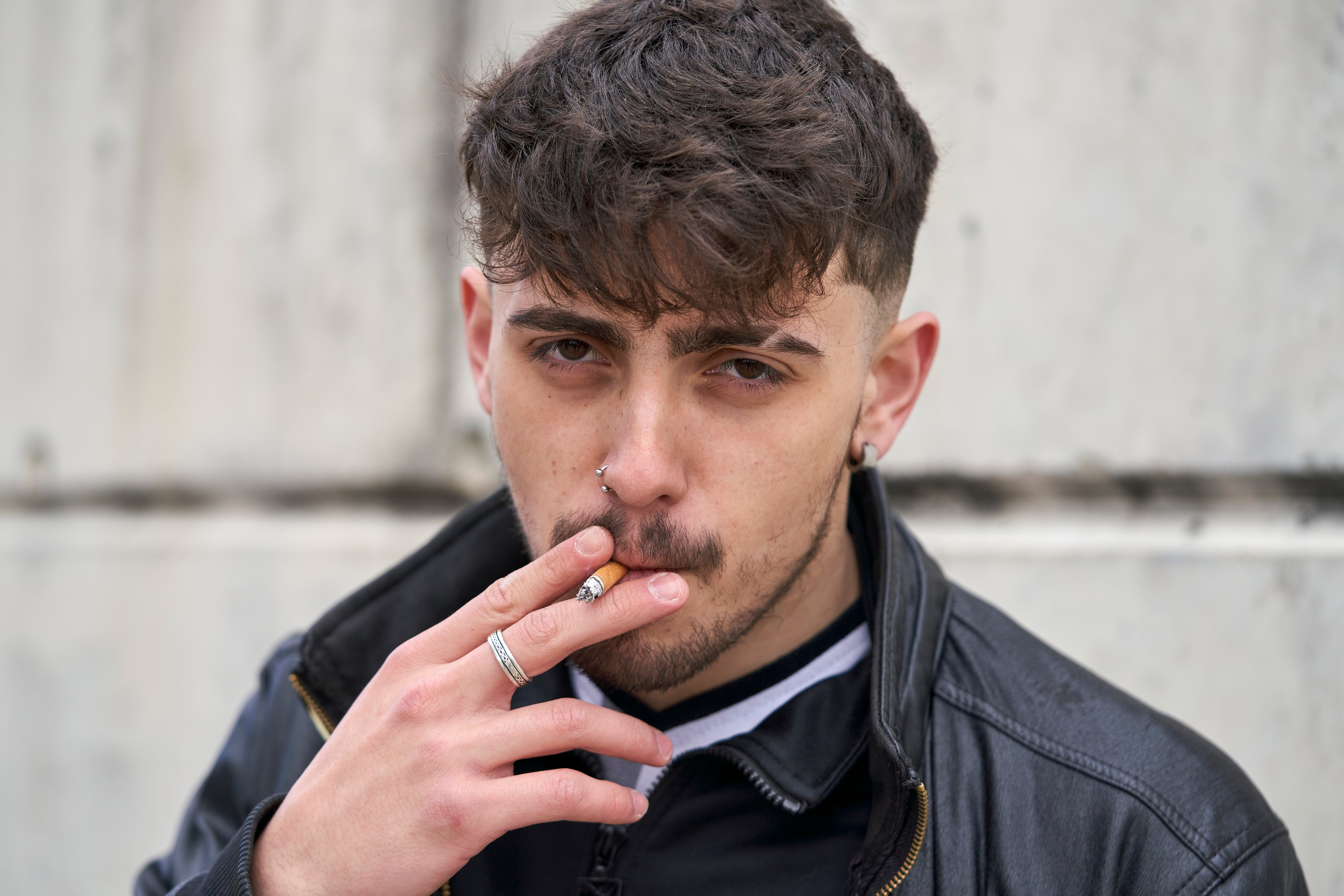 A idiosyncratic with abbreviated hairsbreadth smokes a cigaret outdoors, wearing a leather jacket, conveying themes related to wellness and manner choices