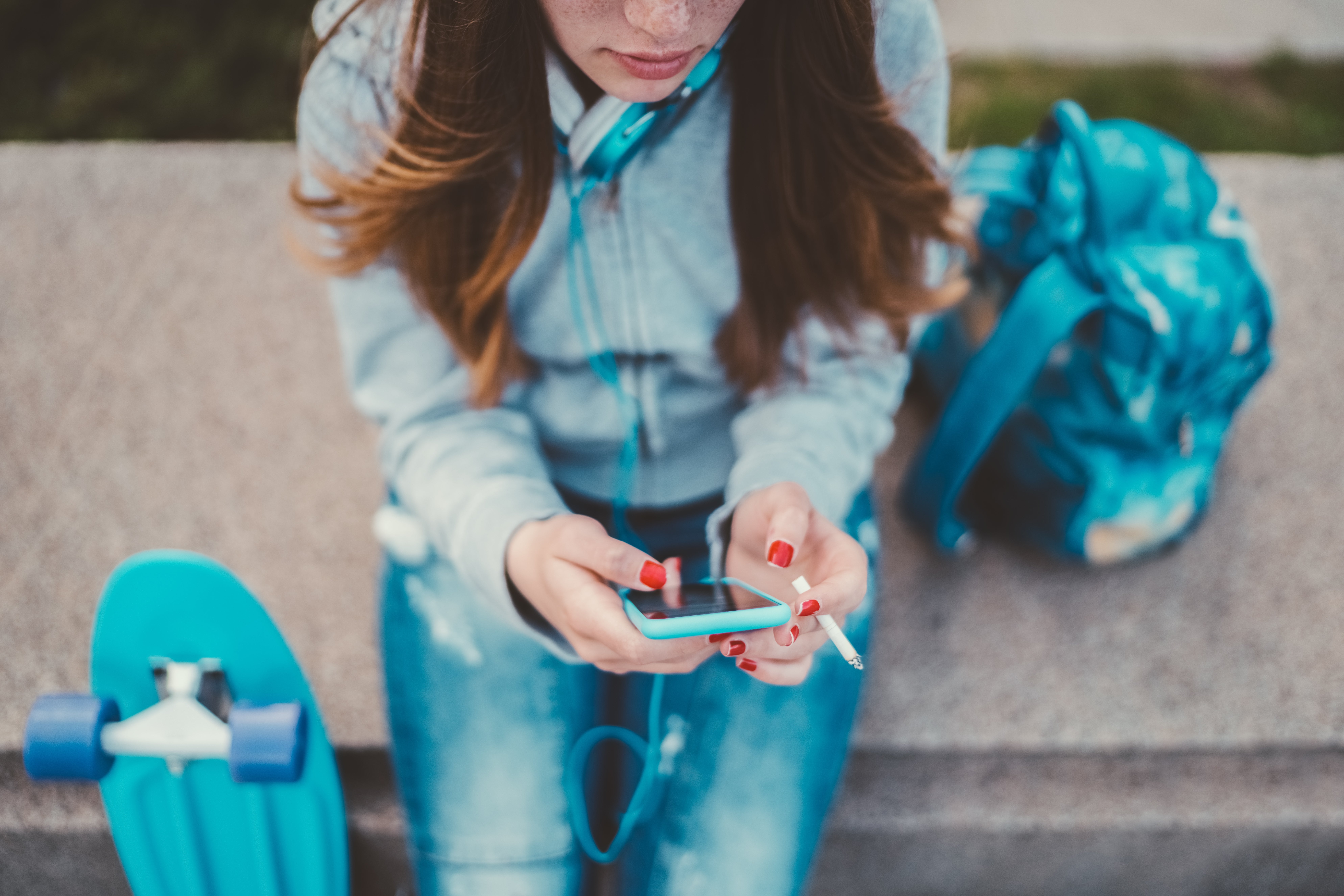Person sitting connected a bench, holding a smartphone and wearing headphones, adjacent a skateboard. Relaxing outdoor leisure moment