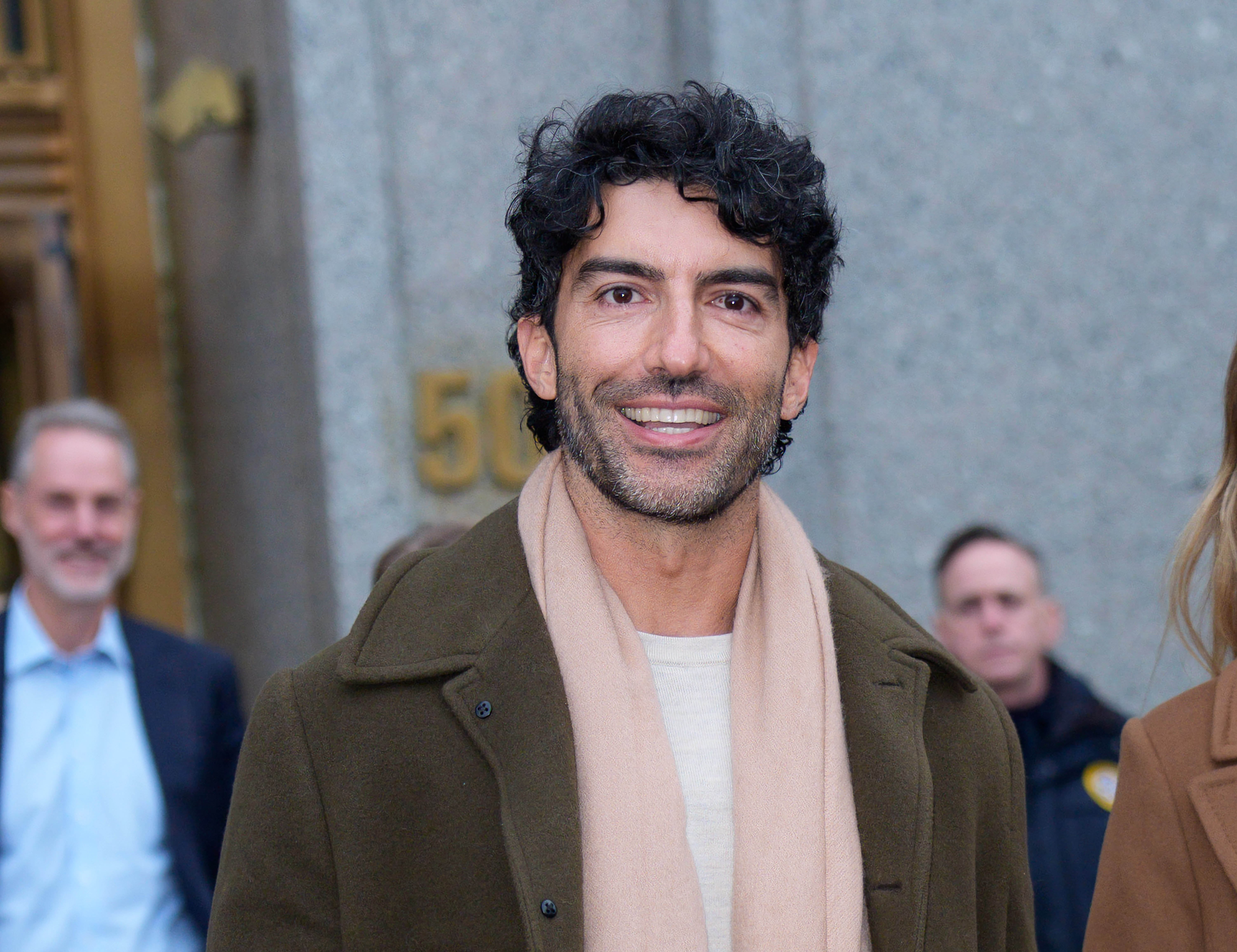 A idiosyncratic   with curly hairsbreadth  smiles extracurricular  a building, wearing a overgarment  and scarf. Others are seen successful  the background