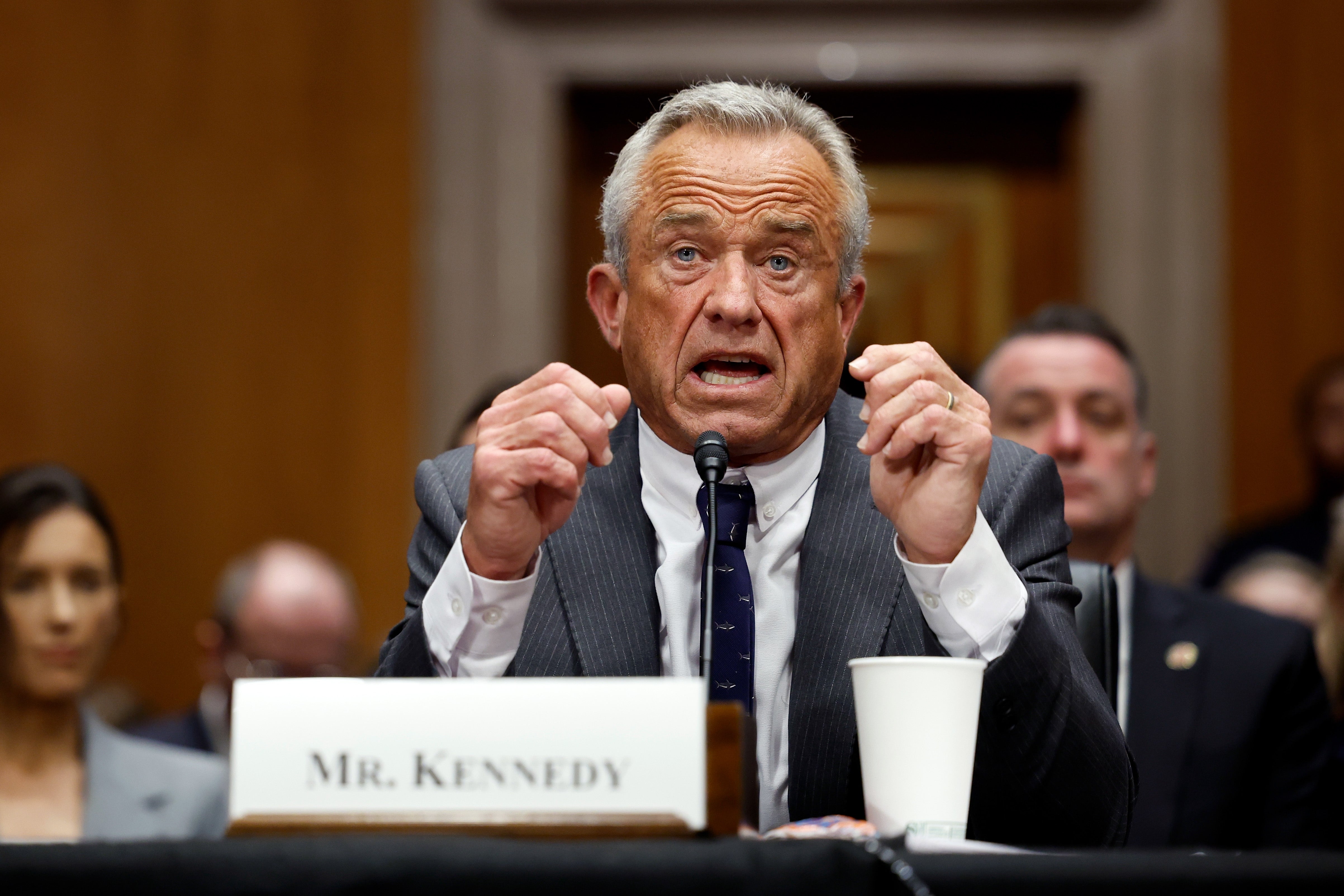 Man successful suit speaking passionately astatine a hearing, seated astatine a array with a nameplate speechmaking "Mr. Kennedy." Others are disposable successful the background