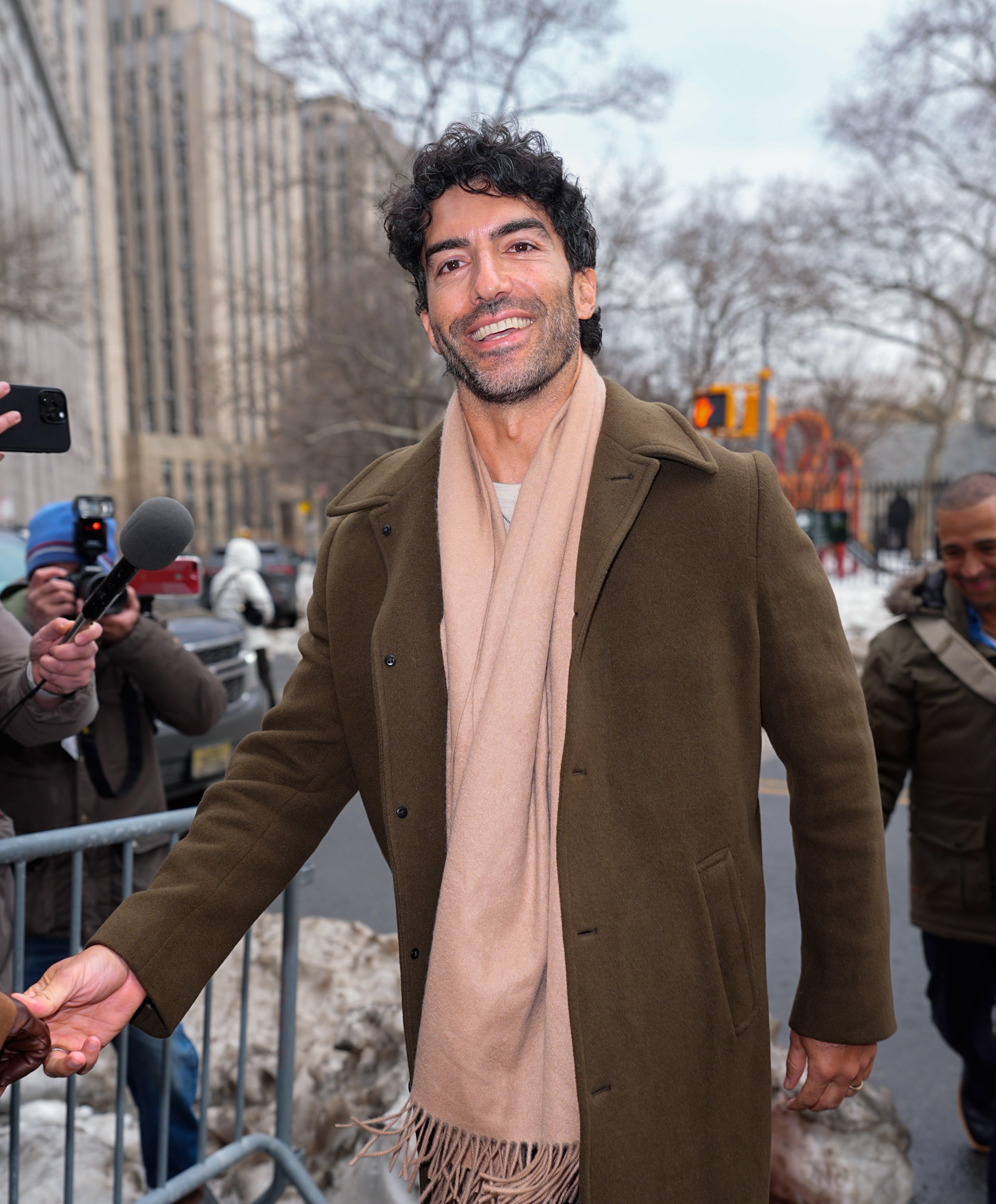 A idiosyncratic   successful  a brownish  overgarment  and beige scarf smiles portion    being photographed outside, with buildings and trees successful  the background