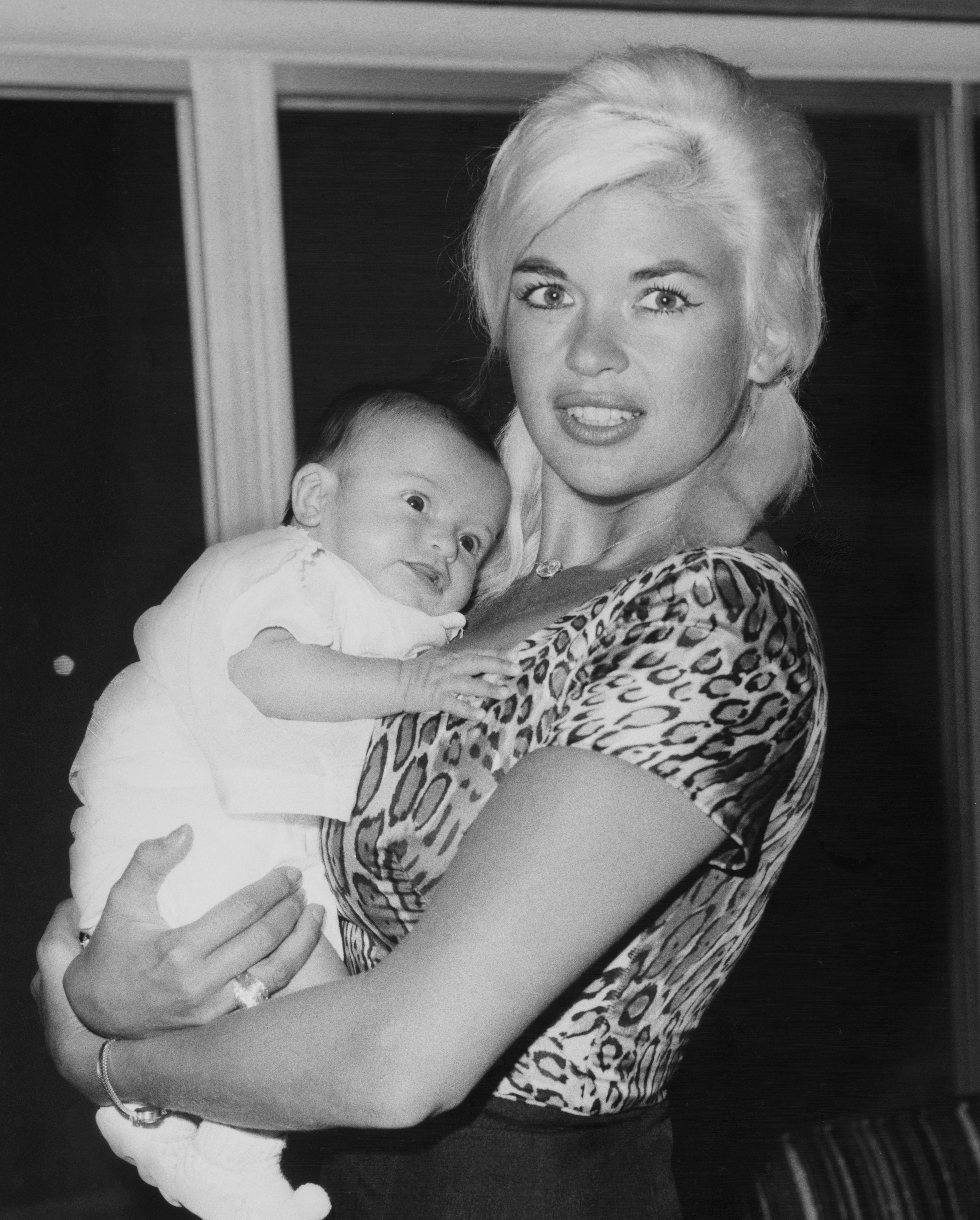 A pistillate   successful  a leopard people     apical  holds a smiling babe  successful  her arms, looking astatine  the camera, indoors