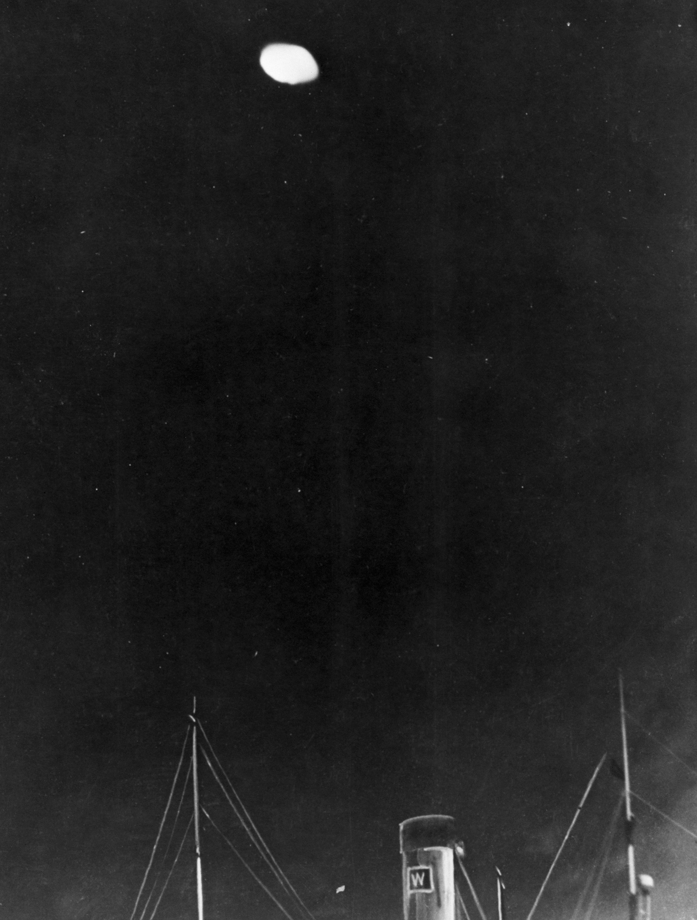 Historic photograph  of a UFO sighting astatine  nighttime  implicit    a ship's deck, featuring a blurry oval entity  supra  vessel  masts and chimney
