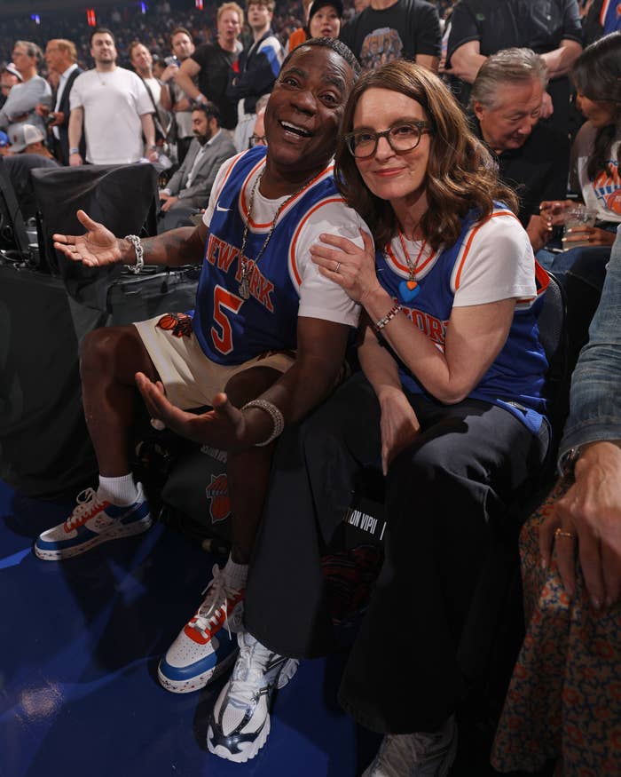 Tracy Morgan and Tina Fey sitting courtside at a basketball game, wearing New York jerseys and smiling at the camera, with a crowd in the background