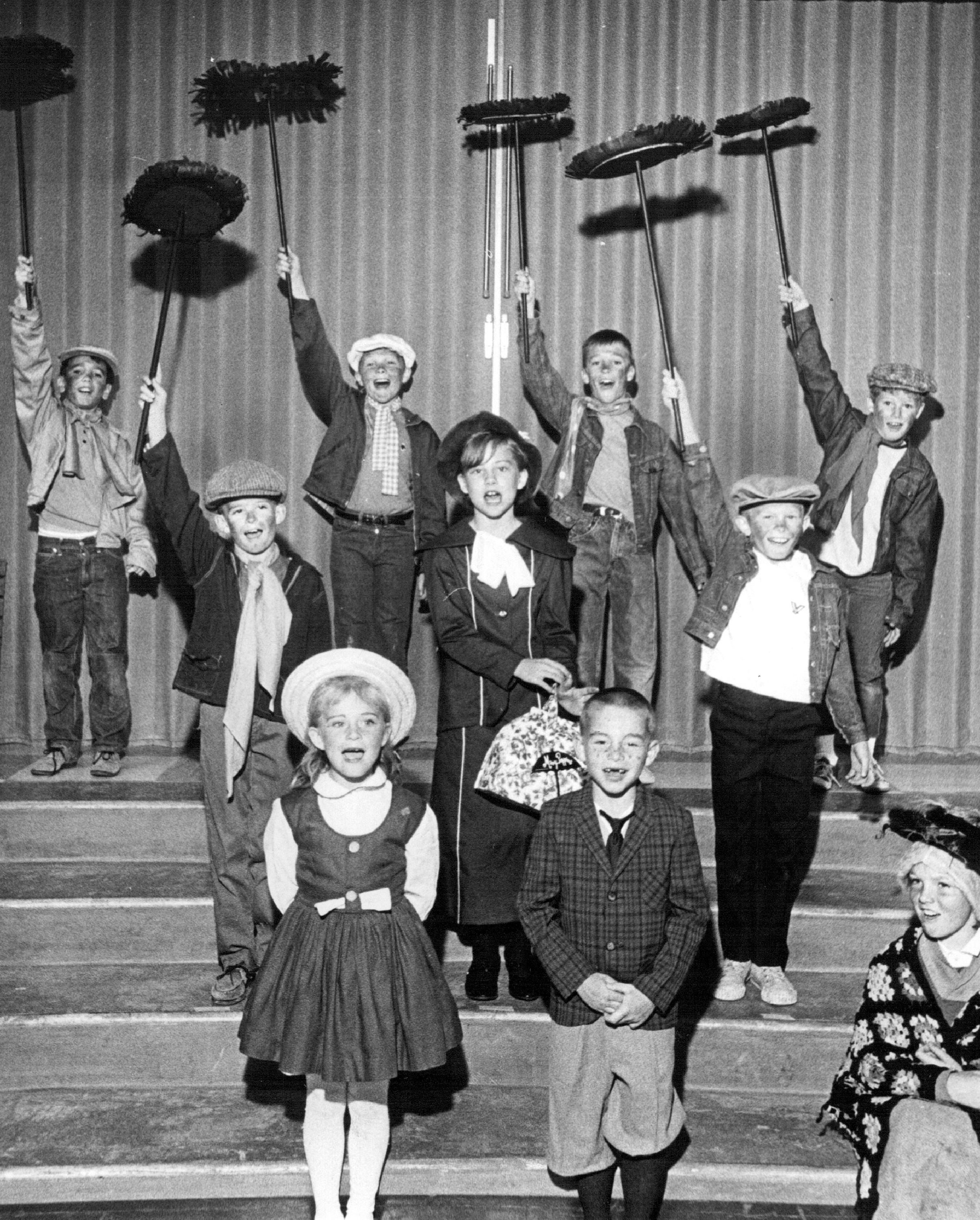 A radical  of children performing a musical, dressed successful  vintage-style costumes, immoderate   holding brooms. They look  to beryllium  successful  a theatrical signifier    setting