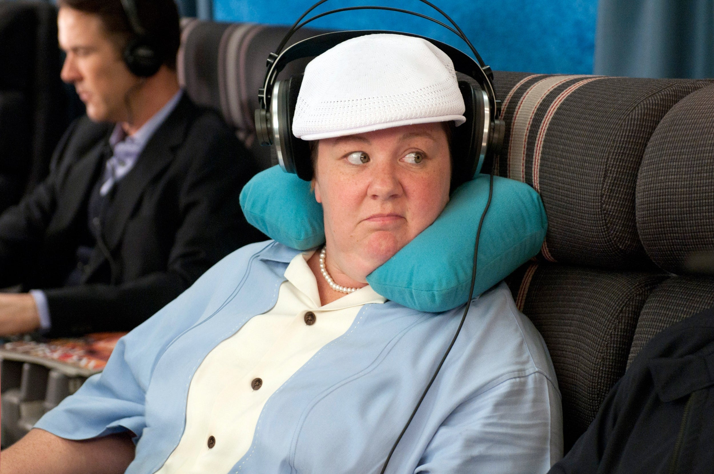 A person wearing headphones and a neck pillow sits on an airplane, looking to the side