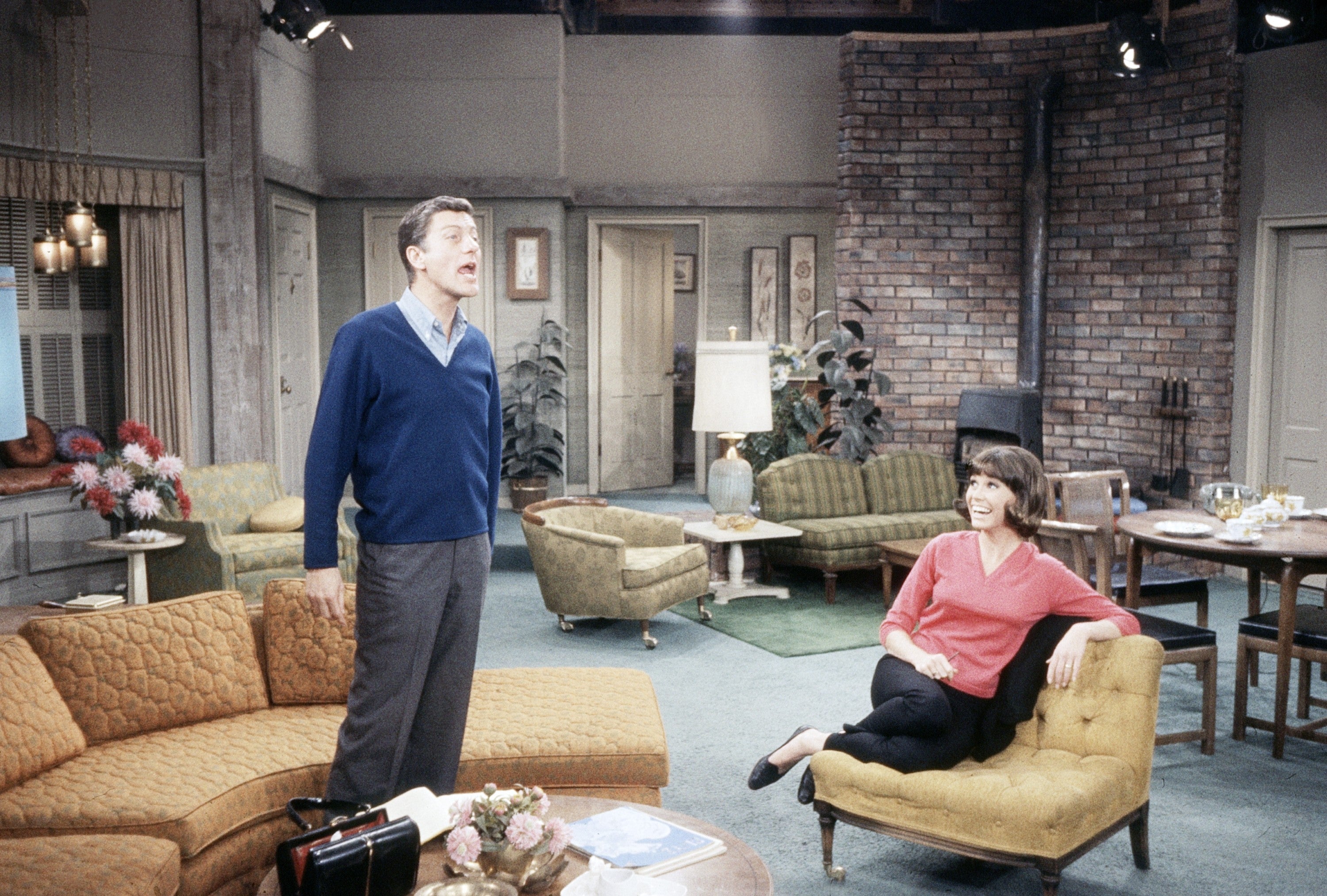 Two actors successful  a retro surviving  country   set; 1  stands animatedly portion    the different   sits laughing connected  a sofa