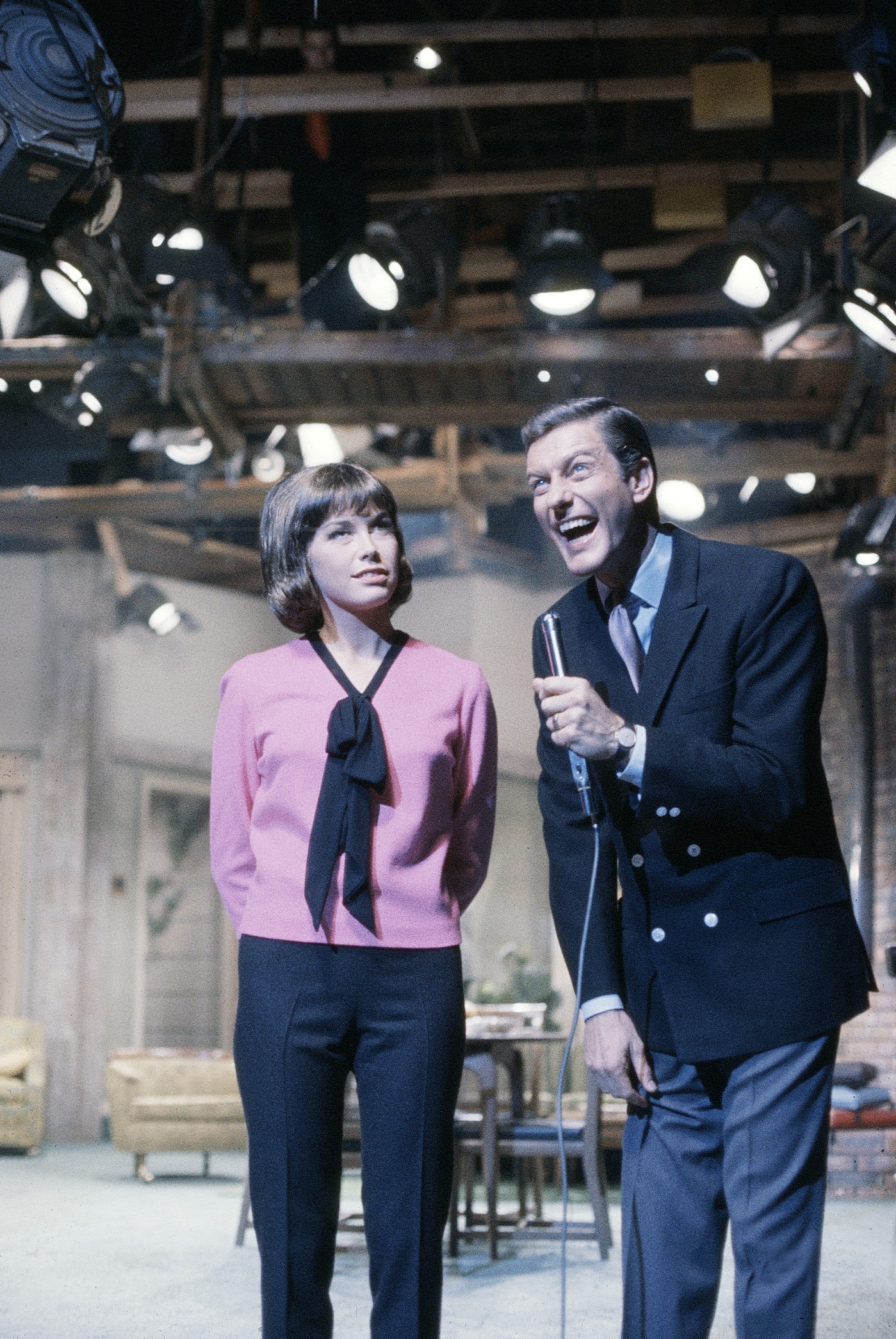A pistillate   successful  a bow-tie blouse stands adjacent  to a antheral   successful  a suit   laughing, some  connected  a TV acceptable   with signifier    lights