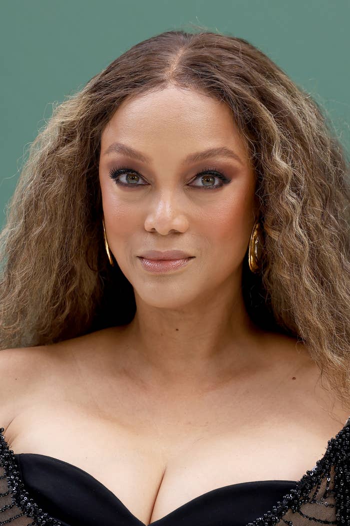 Tyra Banks with long hair and gold earrings poses for a headshot, wearing a stylish off-shoulder outfit with intricate details