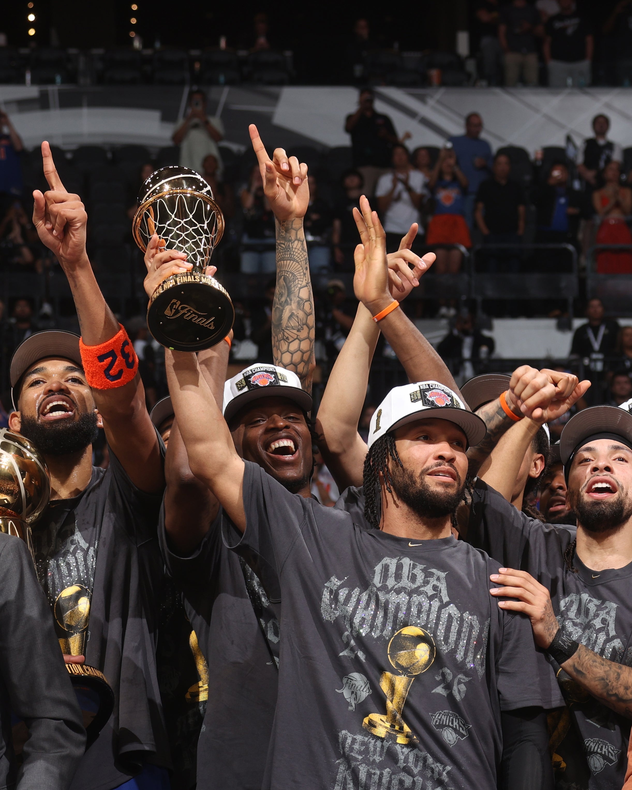 Basketball squad celebrates with trophies and smiles, wearing title shirts and hats, raising fingers successful victory